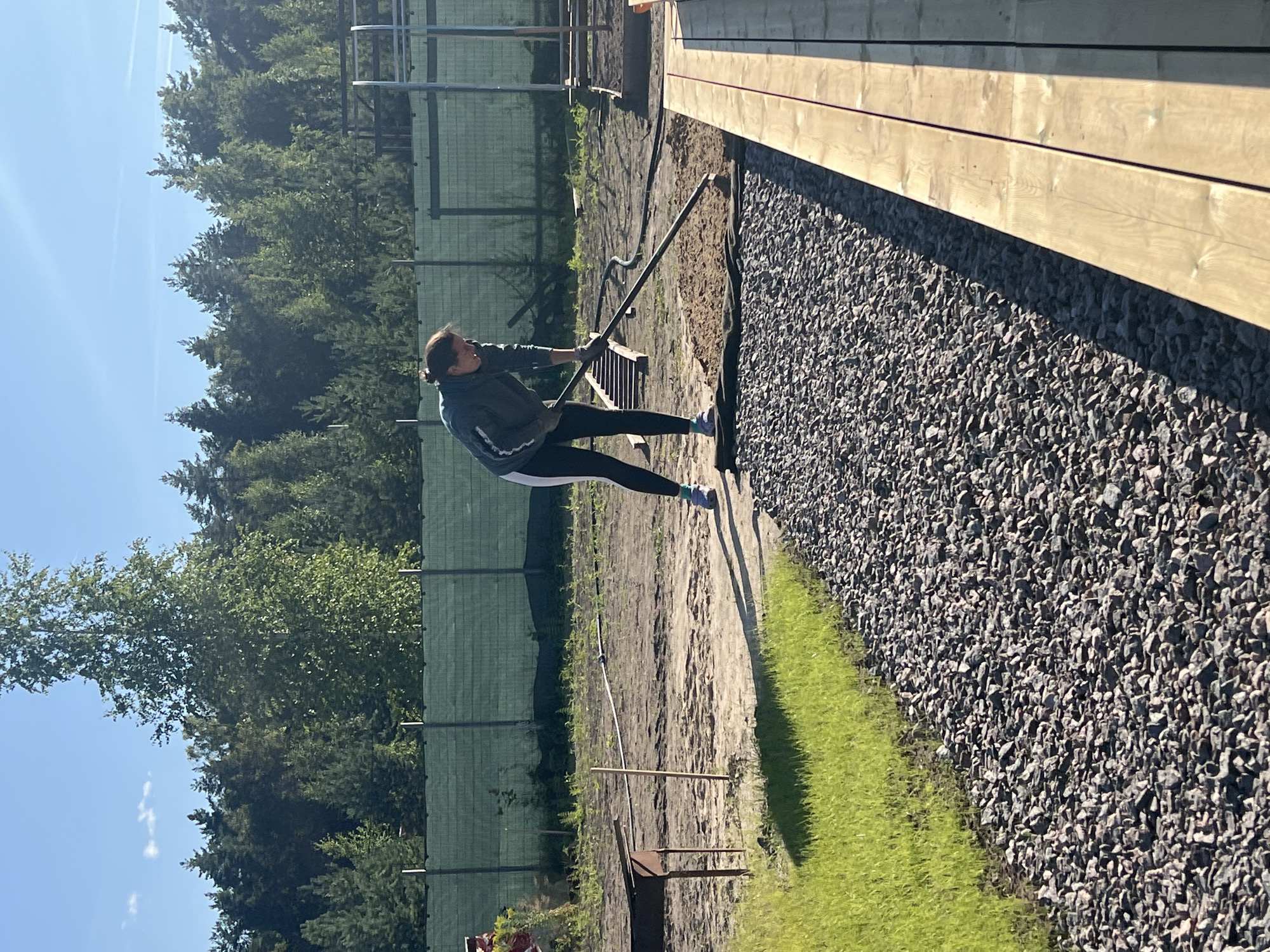 A woman with dark hair tied back is raking brown dirt on a sunny day in a yard. She is wearing a dark grey hooded jacket with white stripes on the sleeves and dark leggings. The woman is standing on a path made of gray gravel, and to her left is a patch of green grass. In the background, there is a green fence, trees, and a blue sky with some white clouds. To the right of the woman, there is a wooden structure.