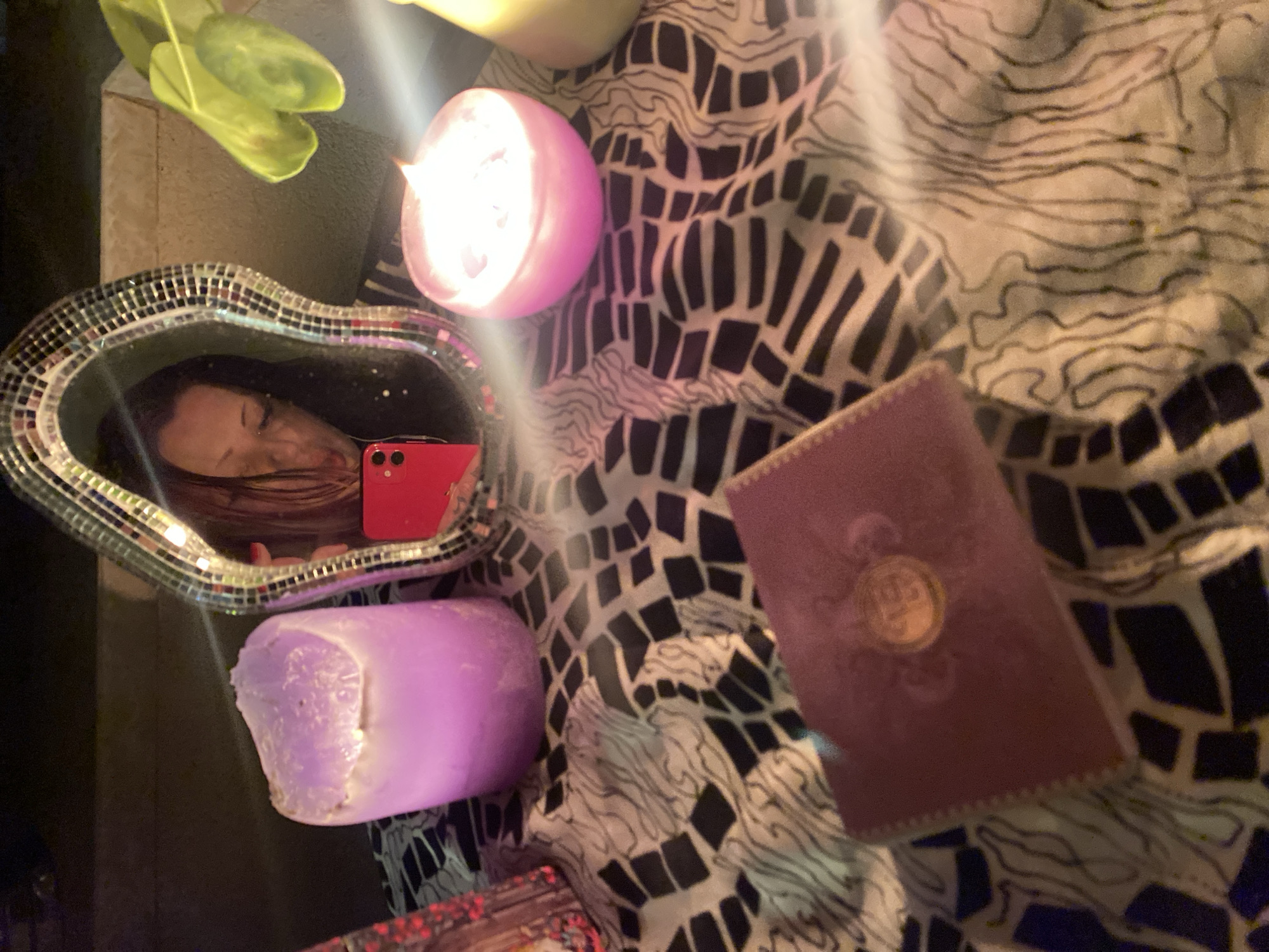 The scene depicts a tabletop arrangement at night in Boreti, Montenegro, illuminated primarily by candlelight. A central, irregularly shaped mirror, framed with mosaic tiles, reflects the upper torso and face of a person with long, dark hair. The person holds a red smartphone, visible in the reflection, positioned for a selfie, with their eyes partially closed and lips pursed. To the left of the mirror is a large, cylindrical lavender candle, partially melted at the top. To the right, a smaller, spherical pink candle is actively burning, emitting a bright flame and casting light. A third, partial, light-colored candle is visible on the far right. In the foreground, a dark pink/maroon rectangular object, resembling a book or box, features a circular gold emblem on its cover. The surface beneath these objects is covered by a fabric displaying a pattern of black geometric shapes and swirling lines on a lighter background. Green plant leaves are visible on the far right, behind the pink candle and against a light-colored wall or surface.