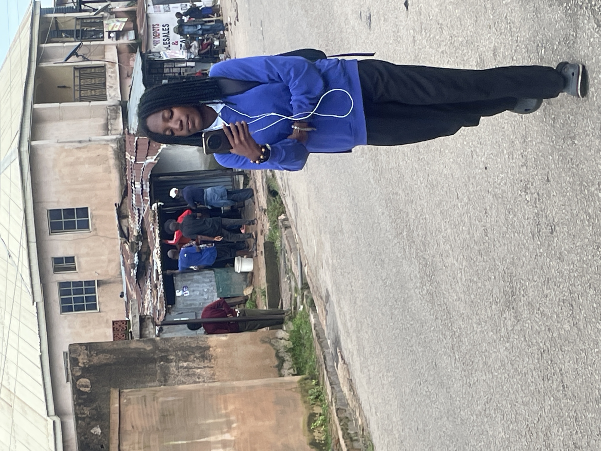 A young Black female walks on a paved road in Jos, Nigeria. She has dark skin, black braided hair, and wears a blue long-sleeved sweatshirt, black pants, and black shoes. She holds a smartphone in her right hand and has white earbuds around her neck. Behind her, several people are gathered near a building with corrugated metal roofing. One person in a red shirt appears to be in conversation with others, while another in a blue shirt stands by a white bucket. The background features a multi-story building with beige walls and windows, and a concrete wall on the left side of the image. The street is lined with debris, and vegetation grows along the curb.