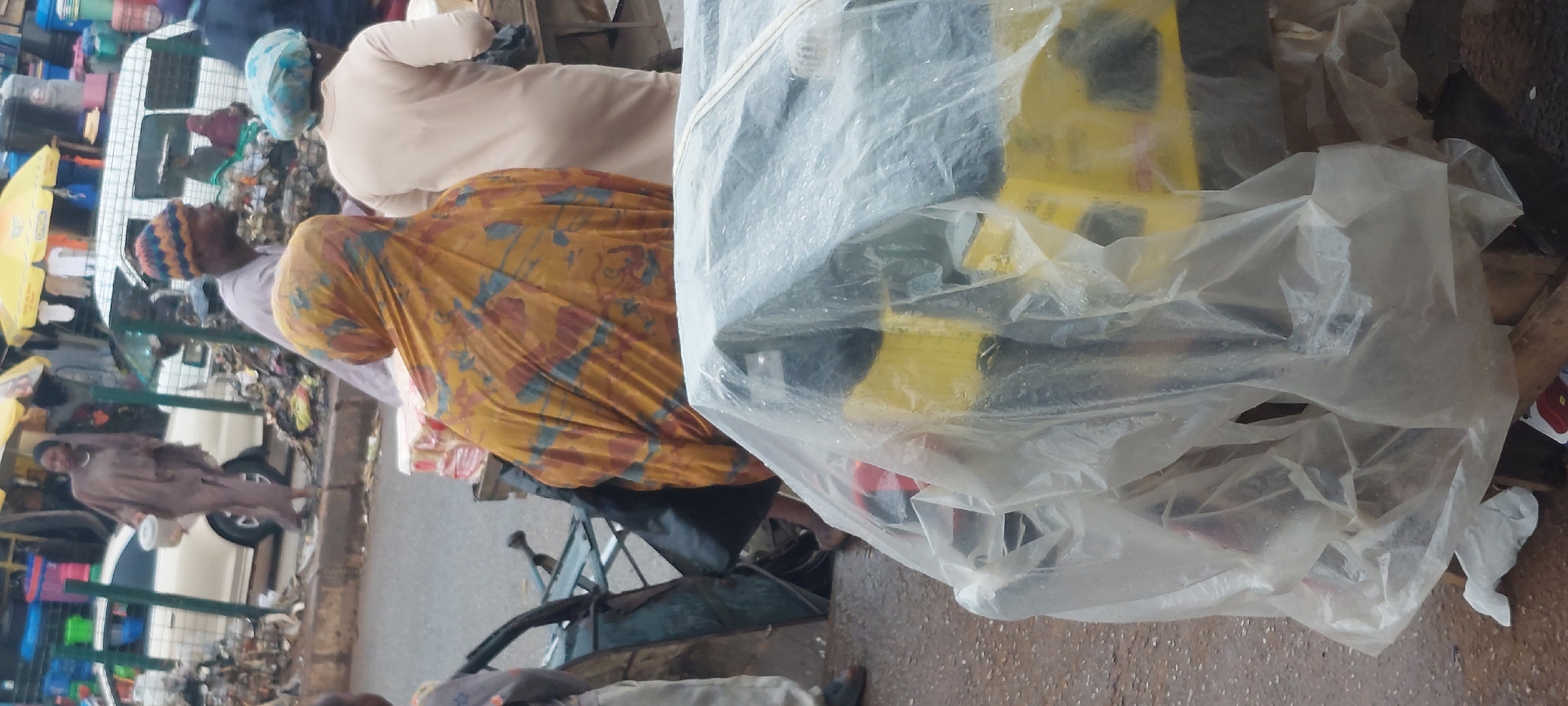 In Bukuru, Nigeria, an outdoor market scene unfolds with a focus on several individuals and goods. In the foreground, a yellow and black object, likely a generator, is partially covered by a clear plastic sheet, suggesting it is either for sale or being protected from the elements. To the left of this object, a metal wheelbarrow with a black bag is visible, indicating active commerce or transportation of goods.
Several people are present. A woman, her back to the camera, wears a vibrantly patterned yellow and red headscarf and dress, possibly engaged in selling or moving merchandise. To her right, another person in a light-colored garment and a blue head covering stands, also facing away. Further back, a man wearing a colorful knitted hat is seen in profile, looking to the right. Behind him, another person in a brown garment walks away, holding a small white container.
The environment appears to be a busy street or market area. Piles of discarded materials and debris line the side of the road. In the background, a white minivan and various stalls with awnings suggest a bustling commercial hub. The ground is wet, indicating recent rain or the general dampness of the area. The overall impression is one of everyday market activity in a Nigerian city.