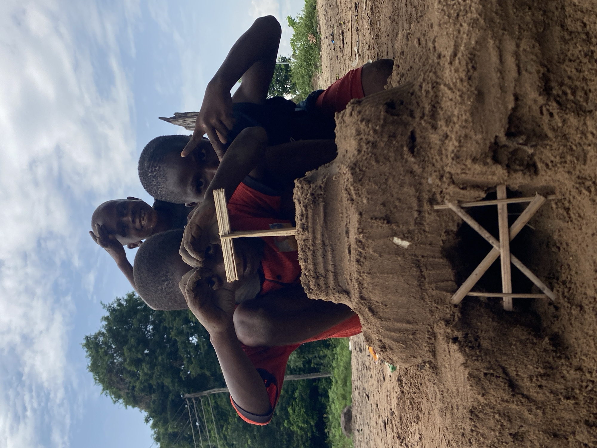 Three African boys pose with a sandcastle structure on the ground in Igbara, Nigeria. The sandcastle has a wooden support structure inside. The boys are touching the structure, smiling, and gesturing. The sky is visible, partially obscured by trees. The ground is sandy.