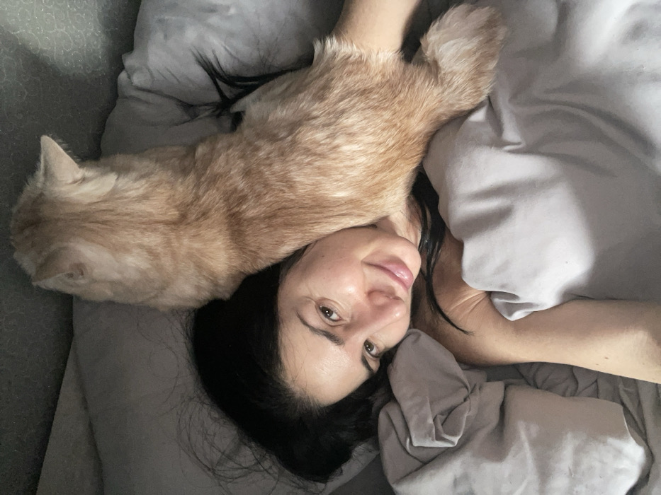 The image displays a close-up view of an individual and a domestic cat in a bed, located in Syasstroy, Russia. A person with dark hair is lying on their back, their head positioned in the lower-left portion of the frame. Their face is turned towards the camera, showing a faint smile. An orange tabby cat is resting on the person's upper torso and neck area, oriented with its back towards the viewer. The cat's head is turned away from the camera, facing the upper-left. Both the person and the cat are partially covered by light gray bedding, which also forms the immediate background.