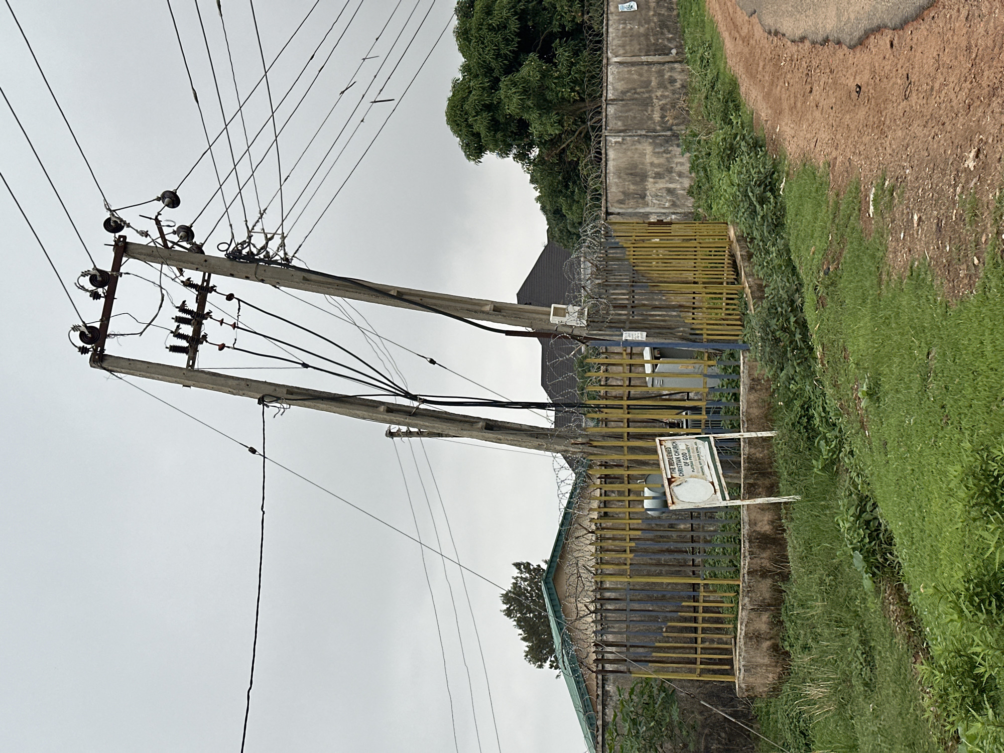 The image depicts an overcast sky over an outdoor scene. In the foreground, a narrow dirt and asphalt road curves to the right. Adjacent to the road is a ditch filled with green grass and some vegetation. Beyond the ditch, a yellow metal fence, topped with barbed wire, encloses an area containing electrical equipment, including a transformer. A sign is visible in front of the fence, reading "THE REDEEMED CHRISTIAN CHURCH OF GOD, PLATEAU PROVINCE". Behind the fence, a building with a green roof is partially visible, along with a concrete wall and more barbed wire. Dominating the center and right of the image are two utility poles with numerous electrical wires extending upwards and across the sky. One pole is tilted at a significant angle. Lush green trees are present in the background. The overall impression is one of an urban or suburban environment with active electrical infrastructure.