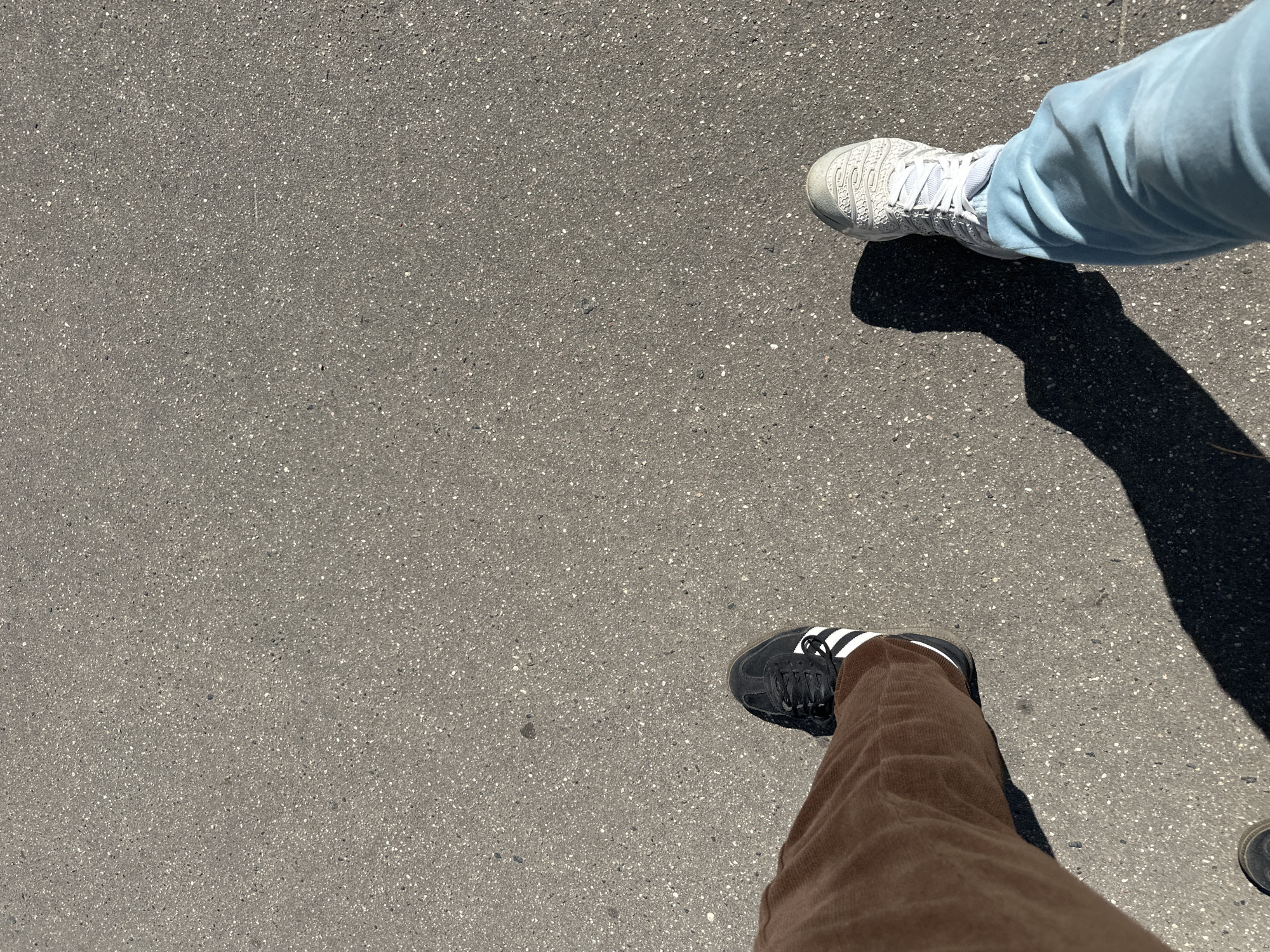 The image shows a first-person perspective of two pairs of feet on an asphalt surface. The left foot is wearing a black Adidas sneaker with white stripes and brown corduroy pants. The right foot is wearing a white sneaker with a textured pattern and light blue pants. Both feet are casting distinct shadows to the right, indicating the sun is to the left. The asphalt appears to be a public walkway or road, with visible aggregate. There are no other people or objects clearly visible in the frame. The scene is devoid of any specific landmarks that would indicate a location in Minsk, Belarus.