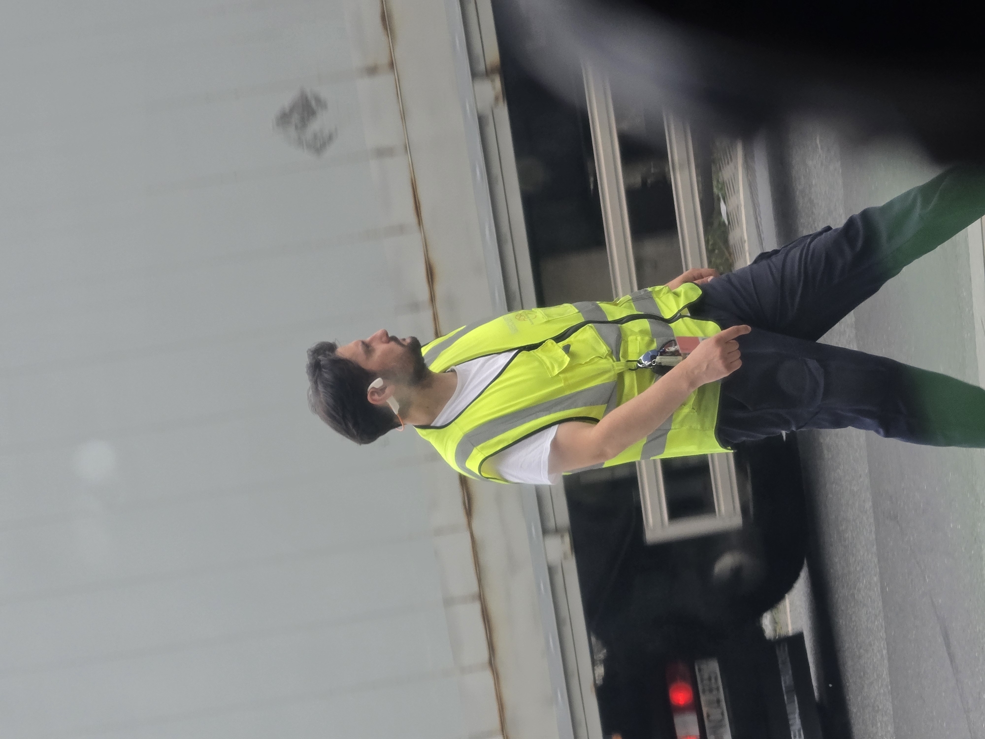 A man wearing a high-visibility yellow vest over a white t-shirt and dark trousers walks across a street in Walldorf, Germany. He has dark hair and a beard and is wearing a white earpiece. A large truck is visible behind him, and a dark blurred object is in the foreground, obscuring part of the scene. The man appears to be moving from right to left in the frame.