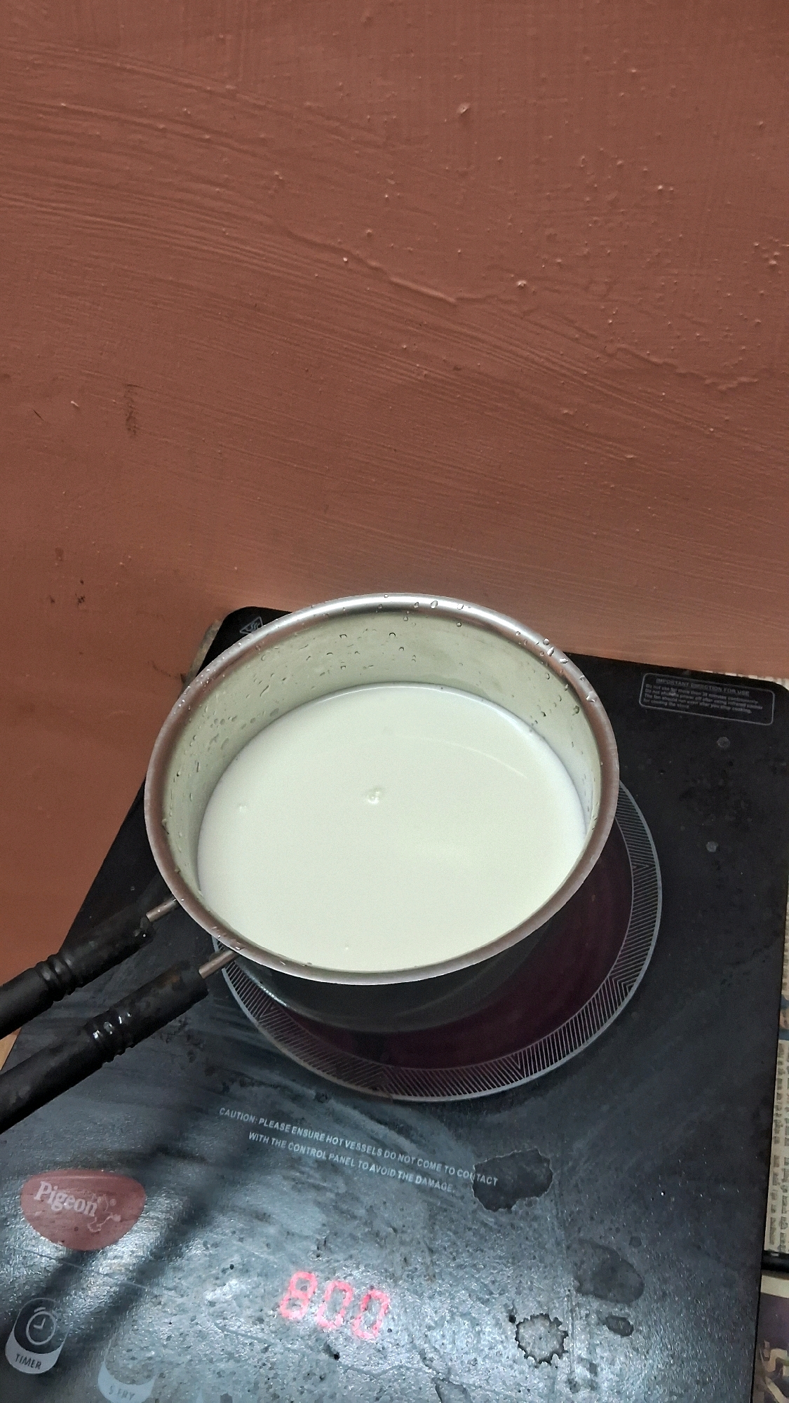 A pot filled with white liquid, possibly milk, sits on a black induction stove. The pot is made of metal and has water droplets on its exterior. The stove has a visible "Pigeon" brand logo and a timer indicator displaying "8:00". The background features a textured, reddish-brown wall. The scene appears to be set in an indoor kitchen.