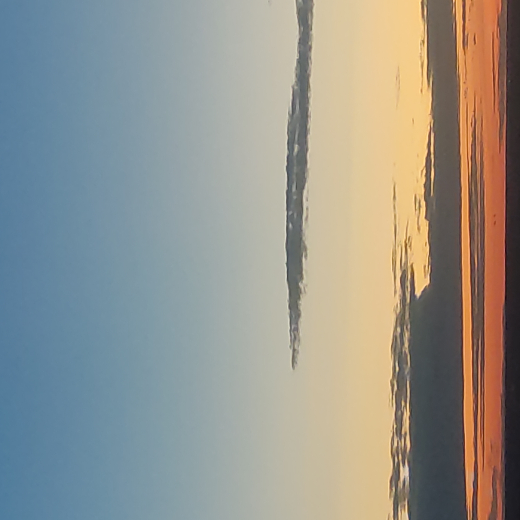 The image shows a sunset or sunrise over Kotsiubynske, Ukraine. The sky transitions from a clear, light blue at the top to yellow and orange hues near the horizon. Several layers of clouds are visible. The lowest layer is dark and dense, illuminated by orange light from below, suggesting it is close to the horizon and catching the direct light of the setting or rising sun. Above this, there are thinner, wispy clouds tinged with orange and yellow. Higher still, a solitary, thin band of clouds stretches horizontally across the sky. The very top of the image is a gradient of pale blue sky. The scene is devoid of any visible people, structures, or distinct geographical landmarks within the frame, making it impossible to infer specific urban details or actions. The focus is entirely on the atmospheric conditions and the interplay of light and clouds.