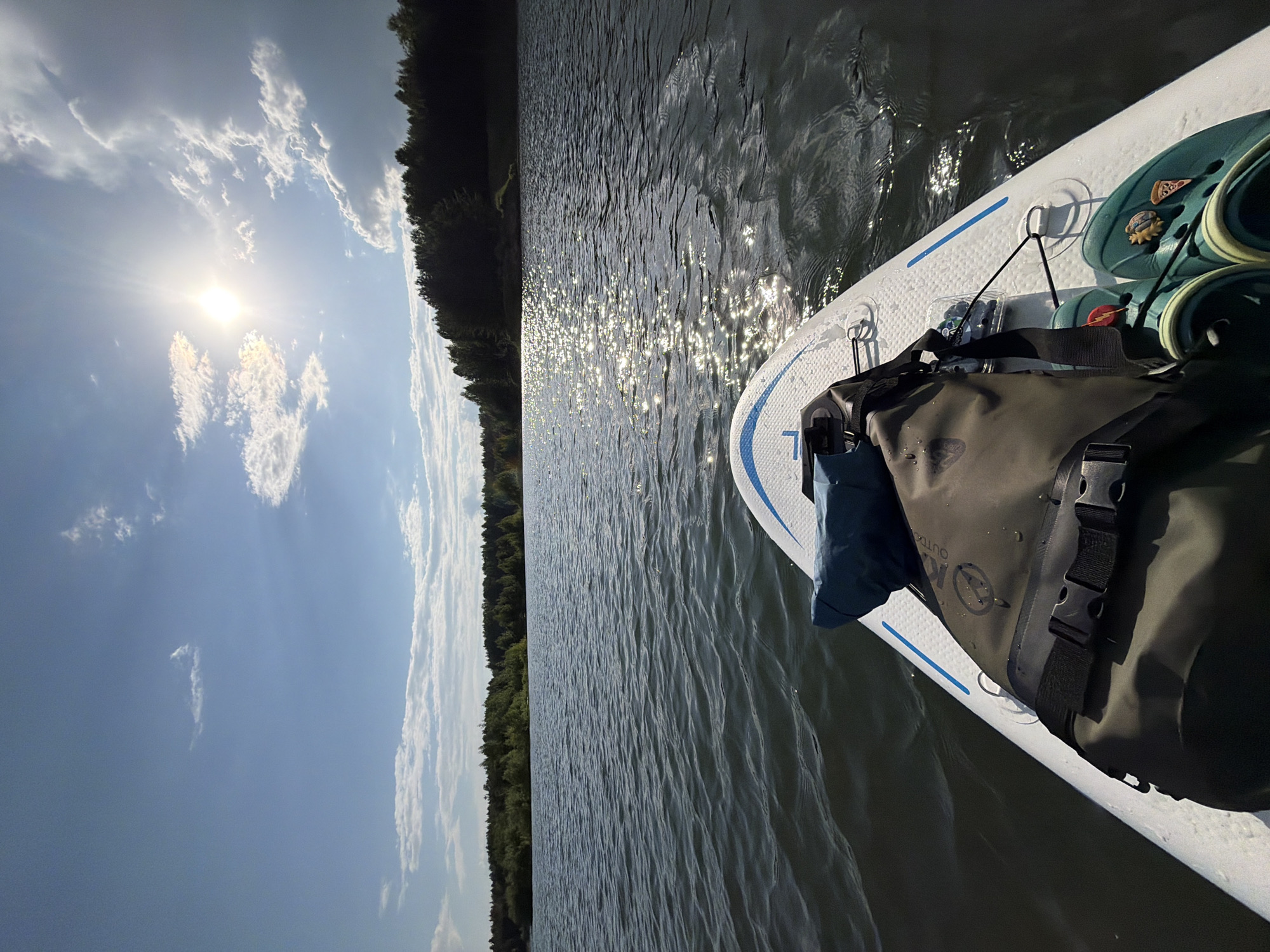 The image shows a sunny day on the water, with a view from a paddleboard. The board is white with blue accents and has a dark olive green dry bag strapped to it. A pair of Crocs, adorned with charms, and a small container, possibly containing berries, are also on the board. The water is shimmering with reflected sunlight, and the opposite shore is lined with a dense forest. The sky is blue with scattered clouds, and the sun is visible. The location is not specified, and therefore the identification of the location in the city of Kanifol'nyi, Russia, is not possible.