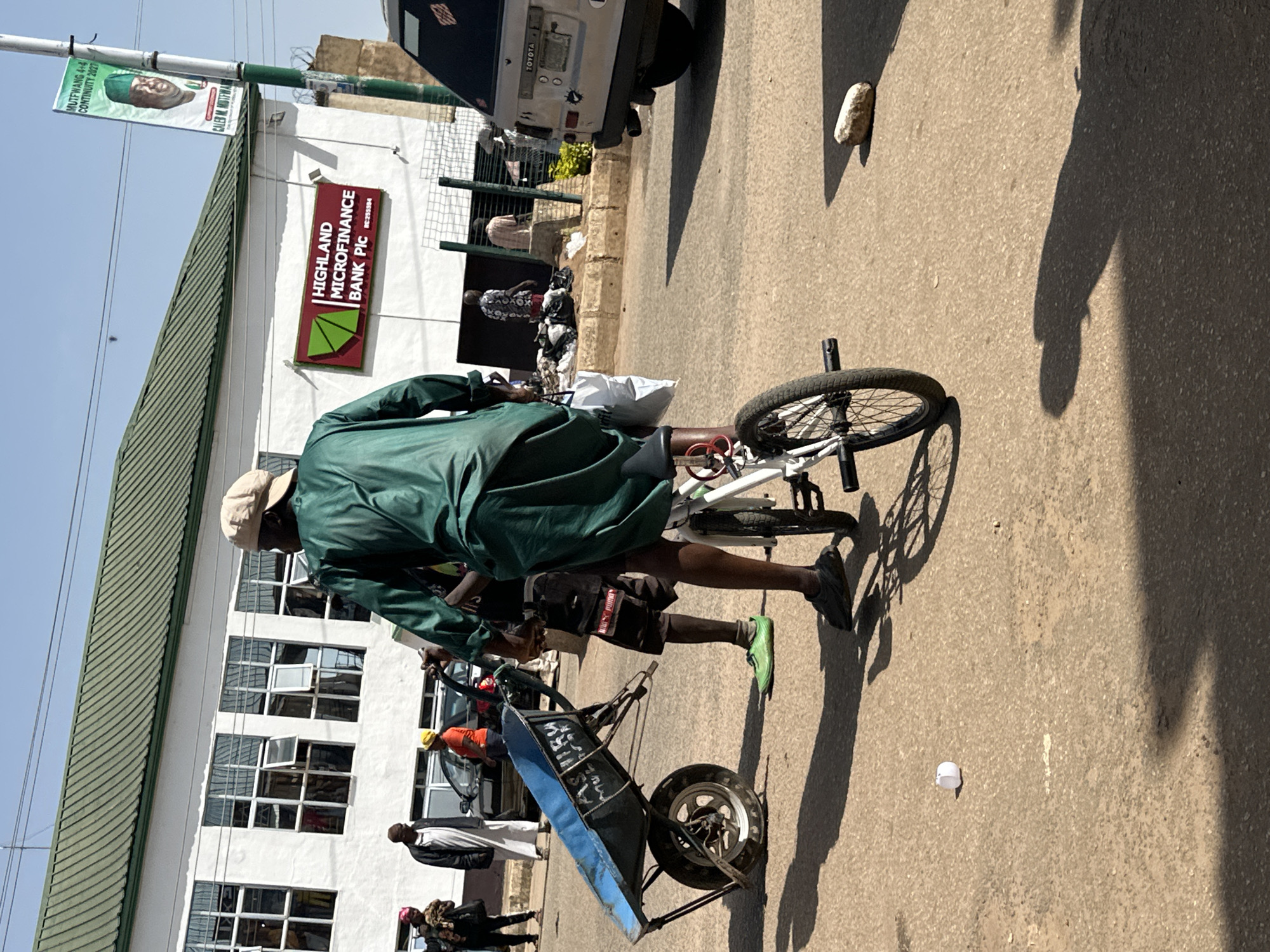 The image depicts a street scene in Bukuru, Nigeria. In the foreground, a man wearing a green robe and a light-colored cap is riding a modified bicycle. The bicycle has a front wheel and a much larger rear wheel, with a frame that appears to be adapted to support a load. To the left of the man, a blue wheelbarrow is positioned on the ground. The wheelbarrow is empty and its handles are resting on the ground. Further in the background, several people are visible. One person is wearing a white robe and head covering, and another is wearing an orange shirt. There is a white building with large windows, bearing a sign that reads "HIGHLAND MICROFINANCE BANK PLC". A banner with a picture of a man and text is visible on a pole to the right of the bank. A white Toyota van is parked on the right side of the road. The ground is paved and appears to be a street or road. The overall scene suggests a typical urban environment in Nigeria.