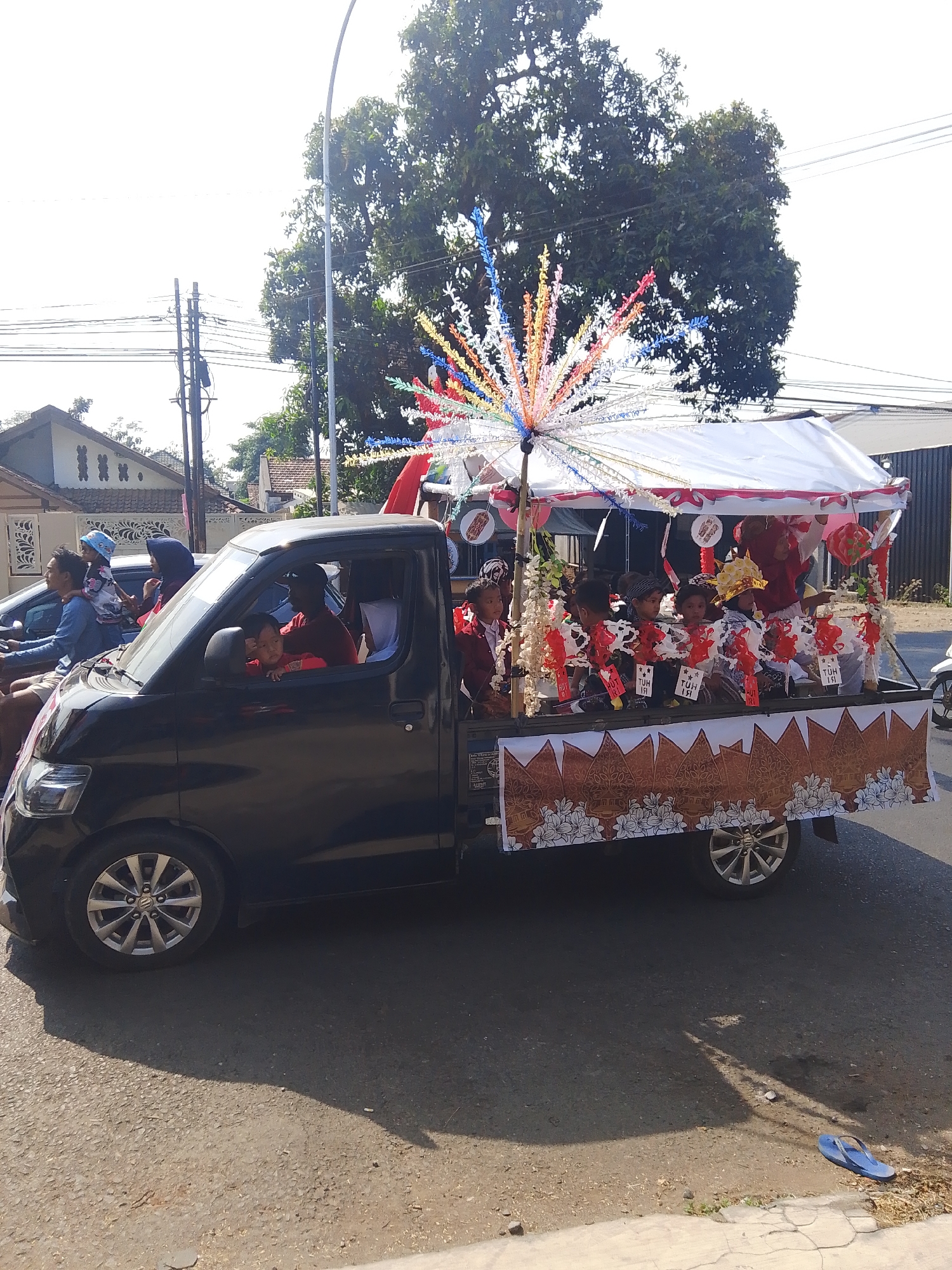 In Kalinyamatan, Indonesia, a black pickup truck with a decorated canopy is driving down a paved street. The truck's bed is adorned with red and white fabric and a banner featuring a traditional Javanese batik pattern. Several children, dressed in festive attire and some wearing hats, are seated in the truck bed, participating in what appears to be a parade or celebration. A colorful, spiky decoration, possibly an umbrella or a float element, is mounted on the canopy. In the driver's cabin, a man is visible, and in the front passenger seat, a child is looking out the window. Other vehicles and people are present on the street, suggesting a lively community event. The background shows houses and trees under a clear sky.