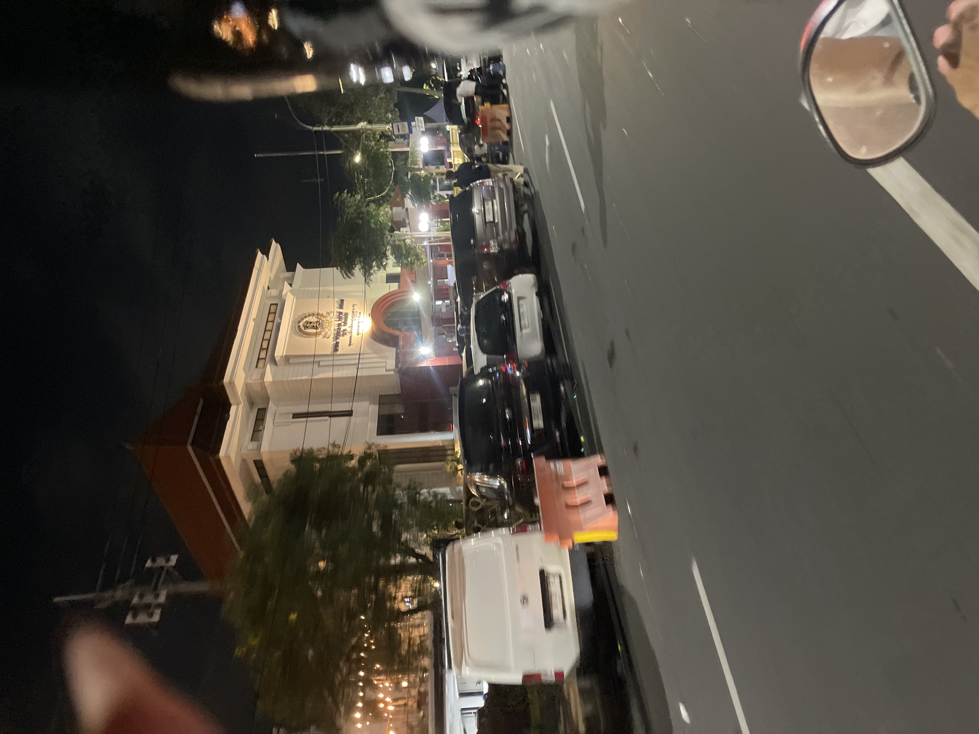 The image captures a night scene on a city street in Surabaya, Indonesia, likely near a government building or institution, indicated by the architecture and signage. In the foreground, a white van is parked, followed by a black SUV, a white hatchback, and a silver SUV. Orange and yellow traffic barriers are visible along the roadside, suggesting some form of road closure or construction. In the background, a multi-story building with a prominent red-tiled roof and detailed facade is illuminated, featuring signage with Indonesian text and a crest. Several streetlights and decorative string lights are also present, casting a warm glow on the scene. People are sparsely visible, some standing near the vehicles and the building entrance. The perspective suggests the image was taken from a moving vehicle, possibly a motorcycle, given the slight blur and the presence of a rearview mirror in the lower right corner. The street itself is paved and appears to be an active thoroughfare, even at night.