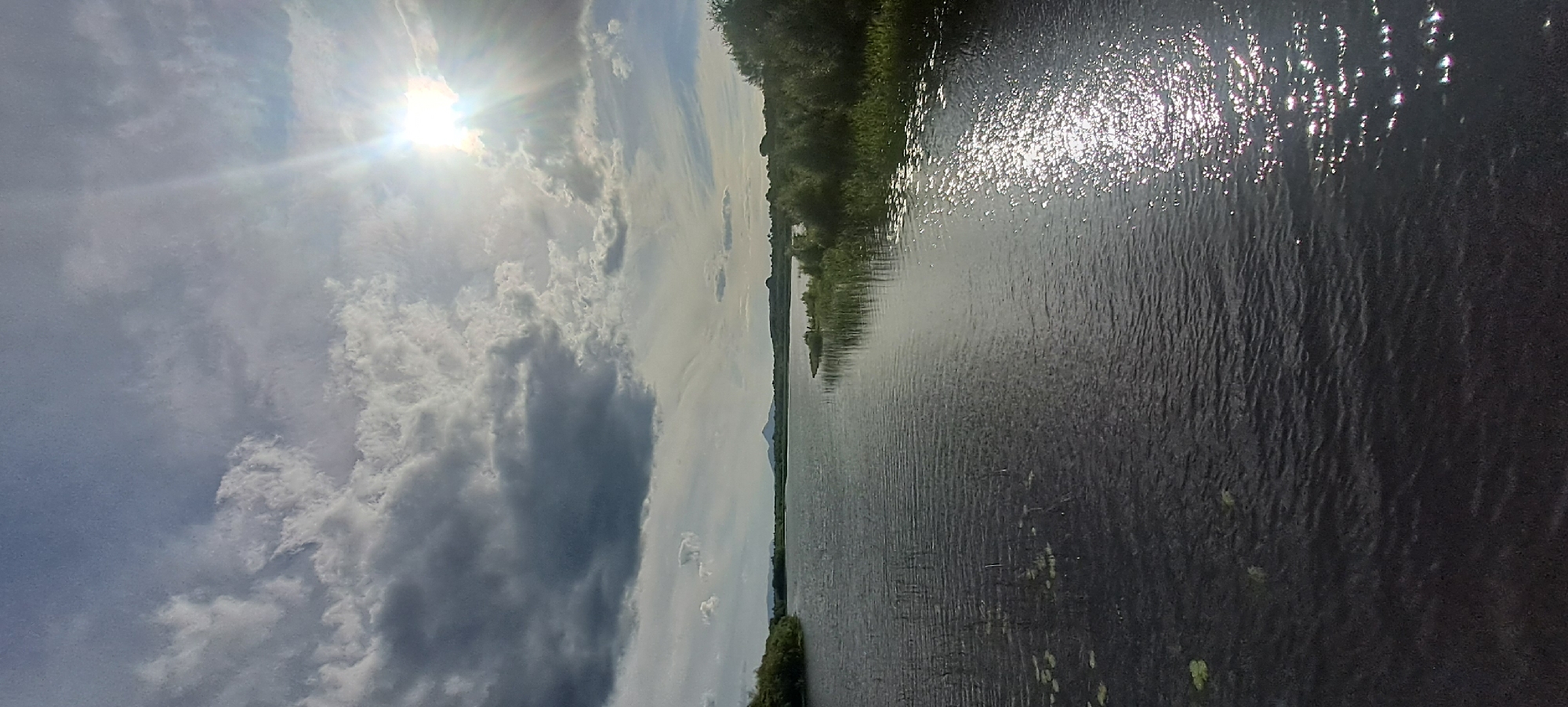 The image shows a wide, dark body of water, likely a lake or river, with ripples on its surface. The sun is shining brightly from the upper right quadrant, casting a strong glare and sparkling reflections on the water. Dark, heavy clouds dominate the sky, with patches of lighter blue sky visible. On the far side of the water, a treeline and low hills can be seen. A mountain or prominent hill is visible in the distance. The foreground of the image is dark and indistinct, suggesting it was taken from a raised vantage point, possibly a bridge or embankment. The overall impression is one of a dramatic, overcast day with intermittent sunshine over a natural landscape. The mention of Castlebar, Ireland suggests this scene is likely located there, possibly at Lough Lannagh, a well-known lake within the town.