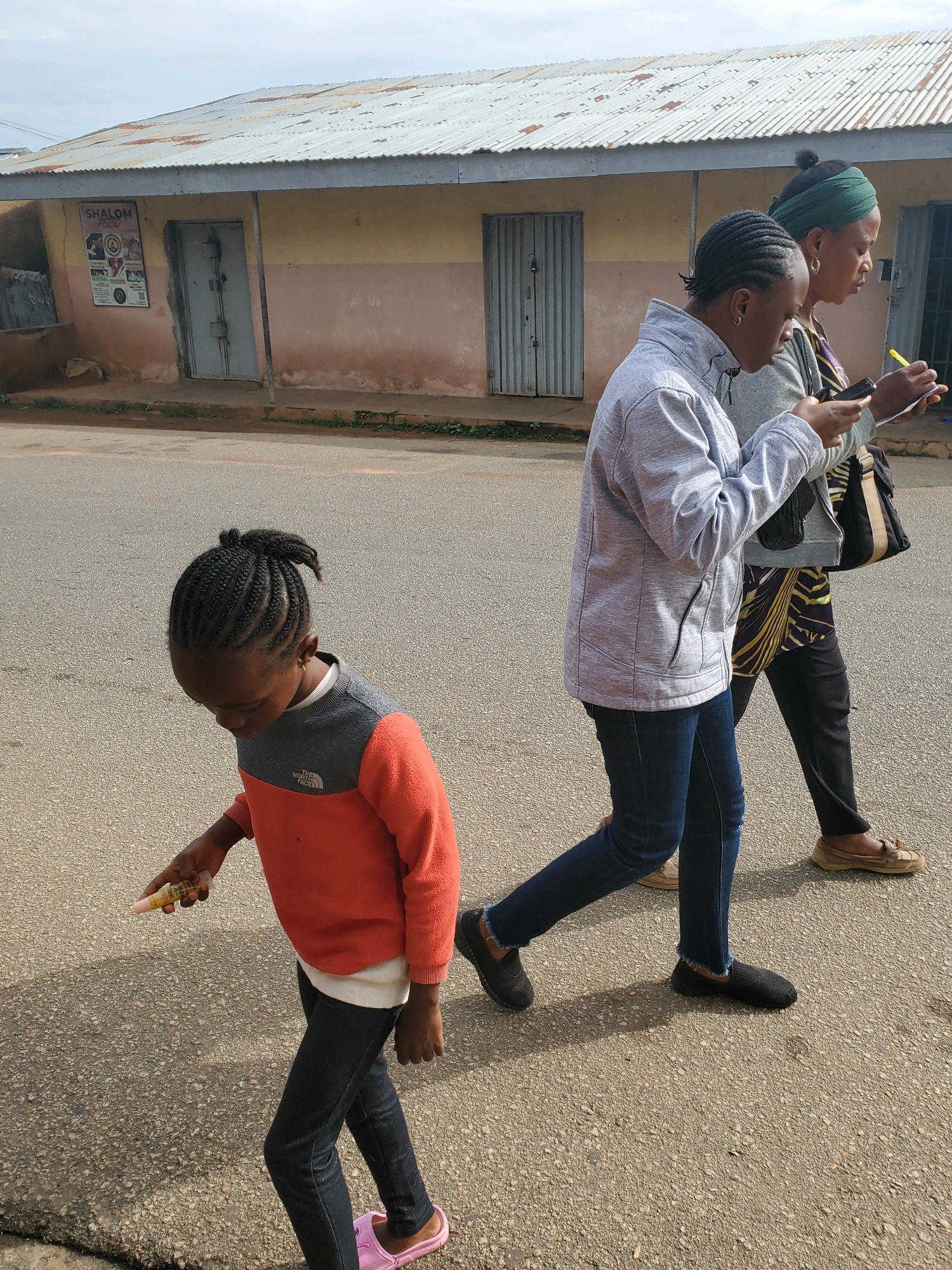 In Jos, Nigeria, a young girl walks down a paved road in front of a building with a corrugated metal roof and a cream and pink facade. She is wearing an orange and grey sweater, dark pants, and pink slip-on shoes. In her right hand, she holds a small tube, possibly lip balm. Behind her, two women walk in the same direction. The woman in the middle wears a grey jacket and dark blue jeans, while the woman on the right wears a dark patterned dress and dark pants. Both women are looking at their phones, and the woman on the right is also holding a yellow pen and a notepad. The building behind them has two large metal doors and a sign that reads "SHALOM FOOD". The ground is a grey asphalt road with some cracks and debris. The sky is overcast.