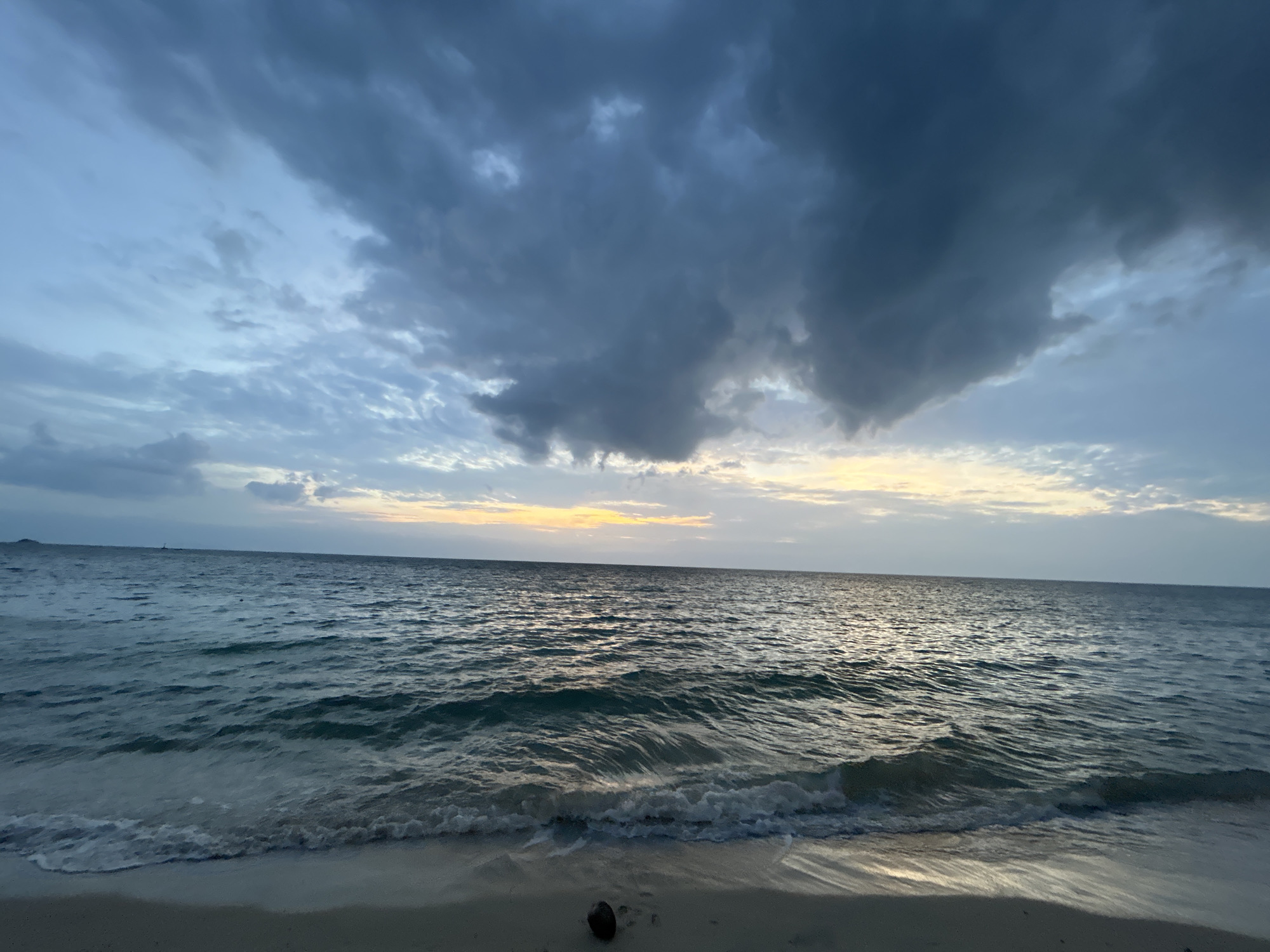 This is a photograph of a beach on Ko Pha-ngan District, Thailand. The image captures a serene beach scene at dusk. The sky is filled with dramatic, dark clouds, but a soft band of yellow and orange light peeks through on the horizon, indicating the setting sun. The ocean is calm, with gentle waves lapping at the shore. The water is a deep blue-green, reflecting the muted colors of the sky. The foreground shows a sandy beach with a few scattered footprints and a single coconut lying near the water's edge. There are no people visible in the image, creating a sense of solitude and tranquility.