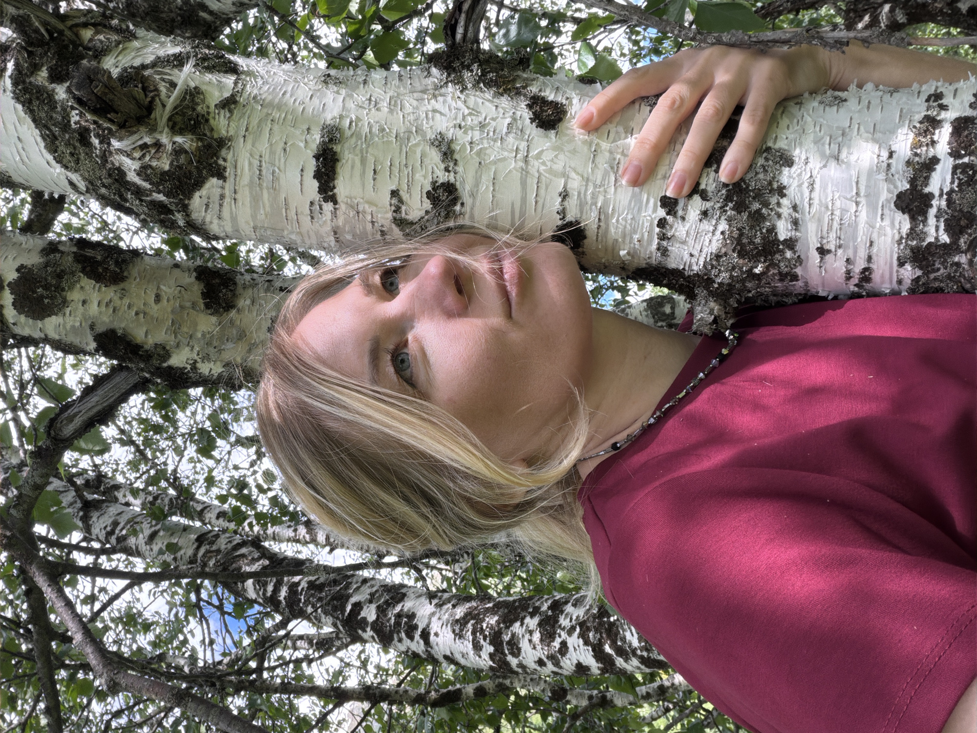 A woman with blonde hair, wearing a maroon t-shirt and a beaded necklace, leans her head against a birch tree in a wooded area. Her right hand rests on the tree trunk. The background consists of more birch trees and foliage, with patches of blue sky visible through the branches. The scene appears to be outdoors, possibly in a park or forest, within the Верхне-Тебердинское сельское поселение, Russia.