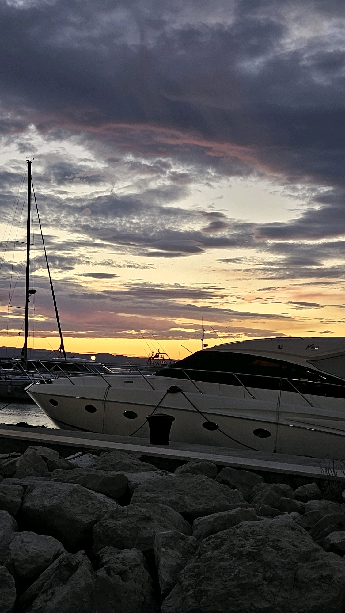 The image depicts a marina in Općina Tučepi, Croatia, at sunset. In the foreground, a rocky shoreline leads to a concrete pier where two yachts are docked. The larger yacht in the foreground is white with black accents and features oval windows along its hull. A black mooring bollard is visible on the pier. In the background, the sky is filled with dramatic clouds painted in shades of purple, pink, and orange as the sun sets behind a distant mountain range. The mast of a sailboat is visible to the left, rising above the yachts. The water is calm, reflecting the colors of the sky.