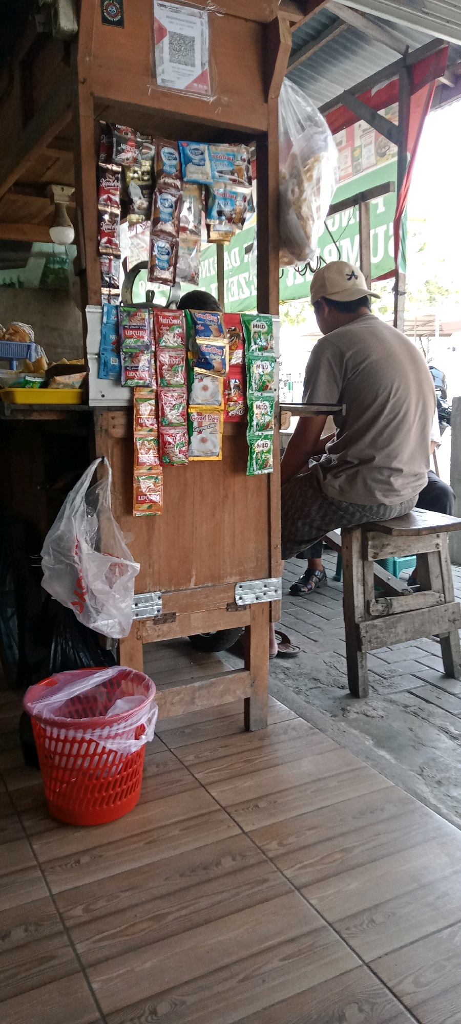 The image depicts a street-side kiosk in Delanggu, Indonesia, where a person is seated on a wooden stool. The kiosk, constructed from wood, displays a variety of packaged goods, including coffee and milk sachets, neatly arranged on its front. A red plastic basket is visible on the tiled floor to the left of the kiosk, containing a plastic bag. A translucent plastic bag with items inside hangs from the kiosk structure. The ground is paved with wood-patterned tiles. The person, wearing a light-colored t-shirt and patterned shorts, is facing away from the camera, looking towards the kiosk. The overall scene suggests a commercial setting, possibly a small local store or a vendor's stall.