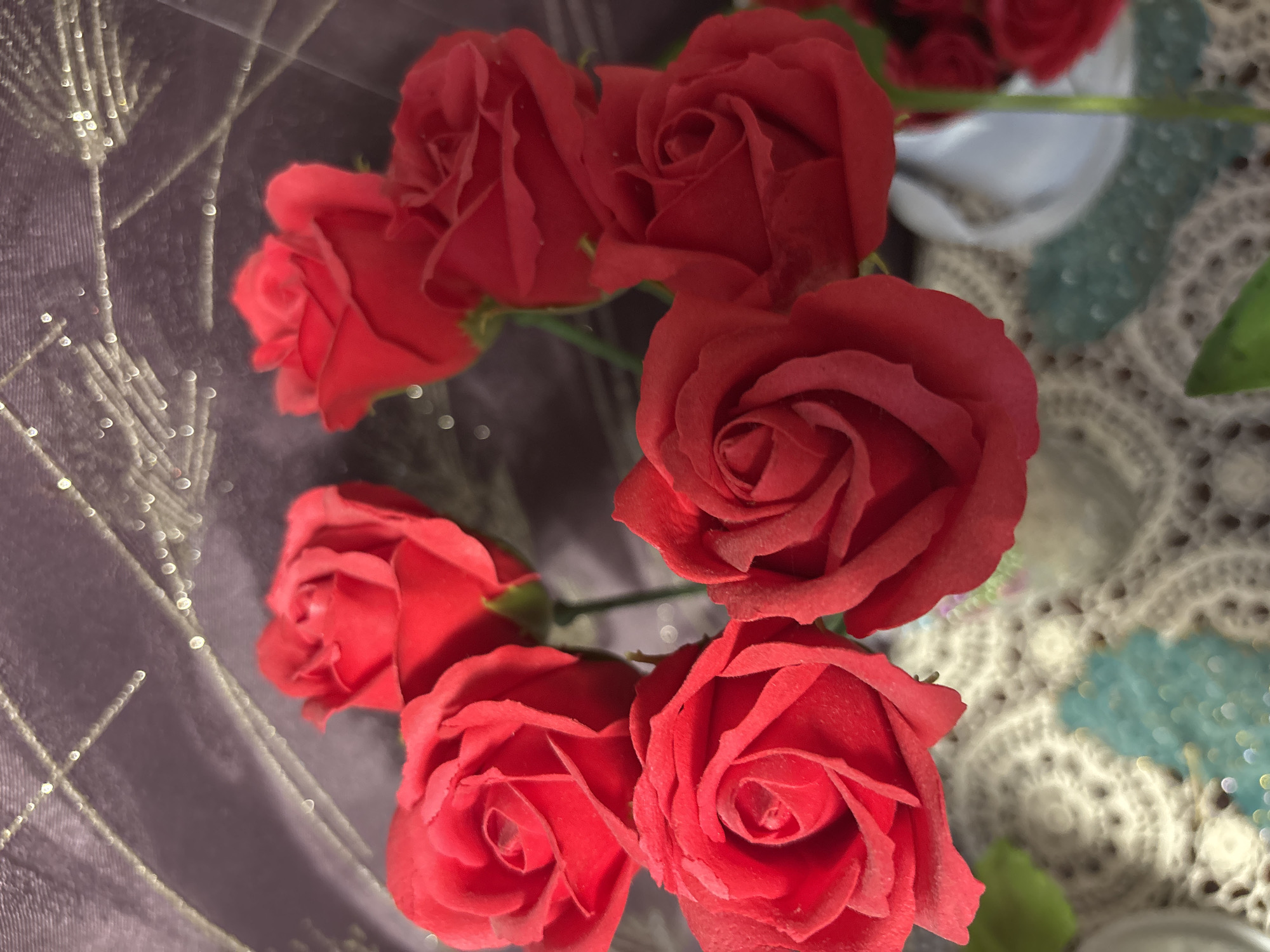 A bouquet of red roses is displayed against a dark purple, textured background with sparkling accents. The roses are arranged in a cluster, with some in sharp focus and others slightly blurred. The foreground shows a glimpse of a doily or lace tablecloth. The setting appears to be indoors.
