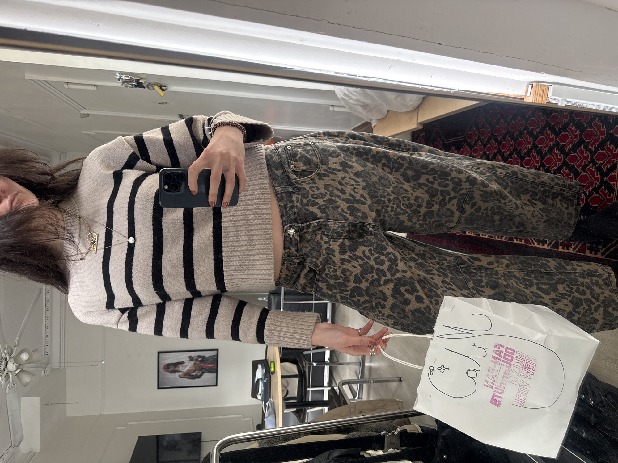 A person is captured in a mirror selfie, showcasing their outfit and a shopping bag in an indoor setting. The image is taken from a slightly upward angle, with the person's reflection visible in a large mirror.
The subject is a female, whose face is partially obscured by her long, dark hair. She wears a beige or light-colored sweater with prominent black horizontal stripes and dark, leopard-print pants. On her left hand, which holds a dark-colored smartphone with multiple camera lenses, her nails are painted a reddish color. A delicate necklace with a small circular pendant is visible around her neck. On her right hand, which holds the white paper shopping bag, she wears several rings. A braided bracelet is visible on her left wrist.
The background reflects a room that appears to be either a retail space or a home, featuring a white ceiling with a ceiling fan or light fixture. A framed picture depicting a person is visible on the wall behind. Furniture, including what looks like a table or desk and chairs, is also reflected. A patterned rug with red and dark motifs can be seen on the floor in the deeper background, while the floor closer to the person appears to be a lighter wood or tile.
The white paper shopping bag held by the person has a stylized drawing and mirrored text. Upon close inspection, the text on the bag reads "FAMOUS DOLLAR PIZZA NEW YORK," along with a distinct, flowing signature-like word that resembles "Calm," and a small flower drawing. The overall scene suggests a casual moment, possibly after a shopping trip in New York, United States of America.