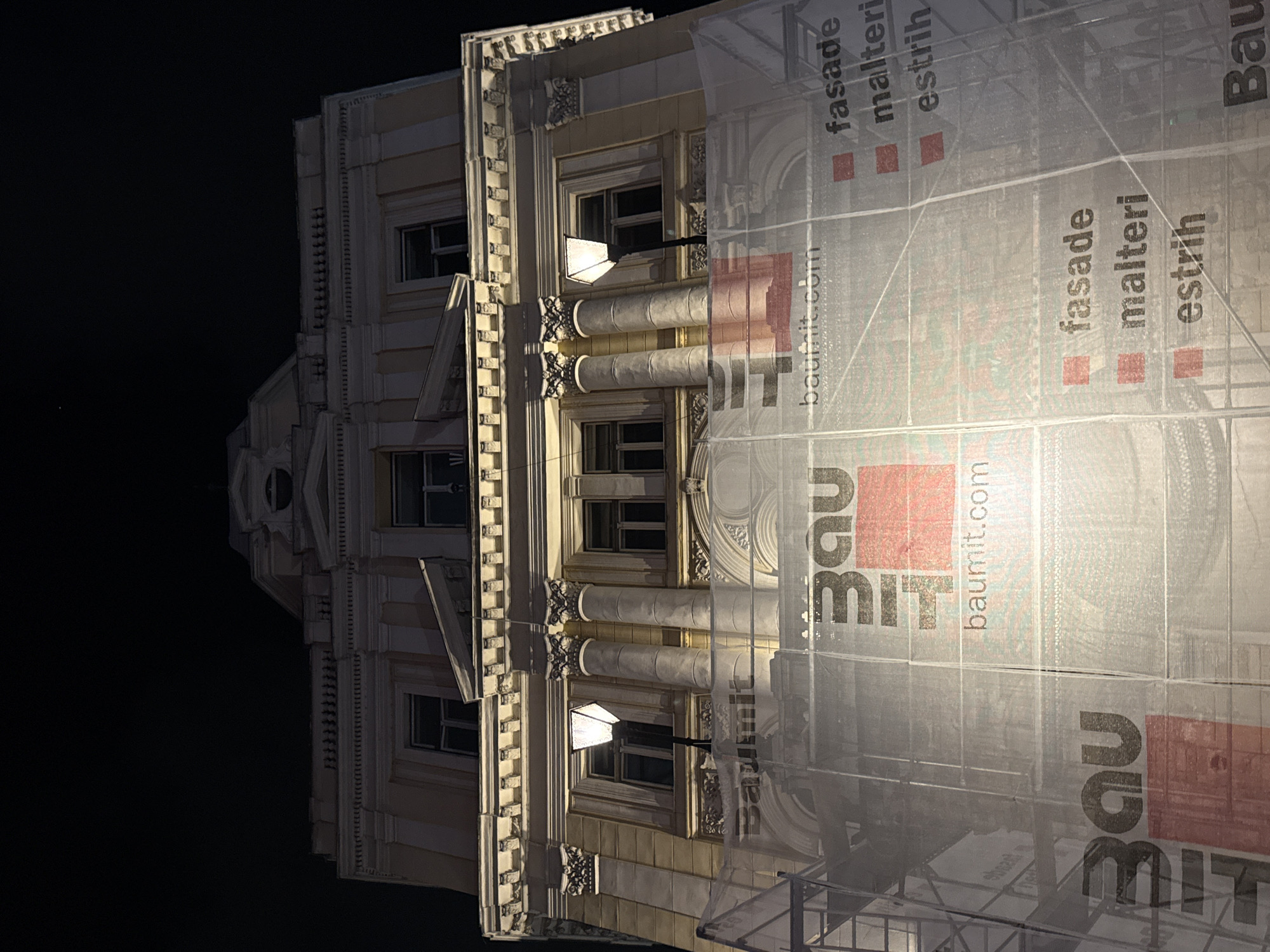 This image captures a nighttime view of a building under renovation in Sarajevo, Bosnia and Herzegovina. The facade of the building, illuminated by artificial lights, features classical architectural elements such as columns and ornate moldings. A significant portion of the lower facade is covered by scaffolding and a white mesh net, which displays branding from "Baumit" and lists building-related services like "fasade," "malteri," and "estrih" in what appears to be Bosnian or a similar South Slavic language. The darkness of the night sky contrasts with the brightly lit building, creating a dramatic effect. No people are visible in the image.