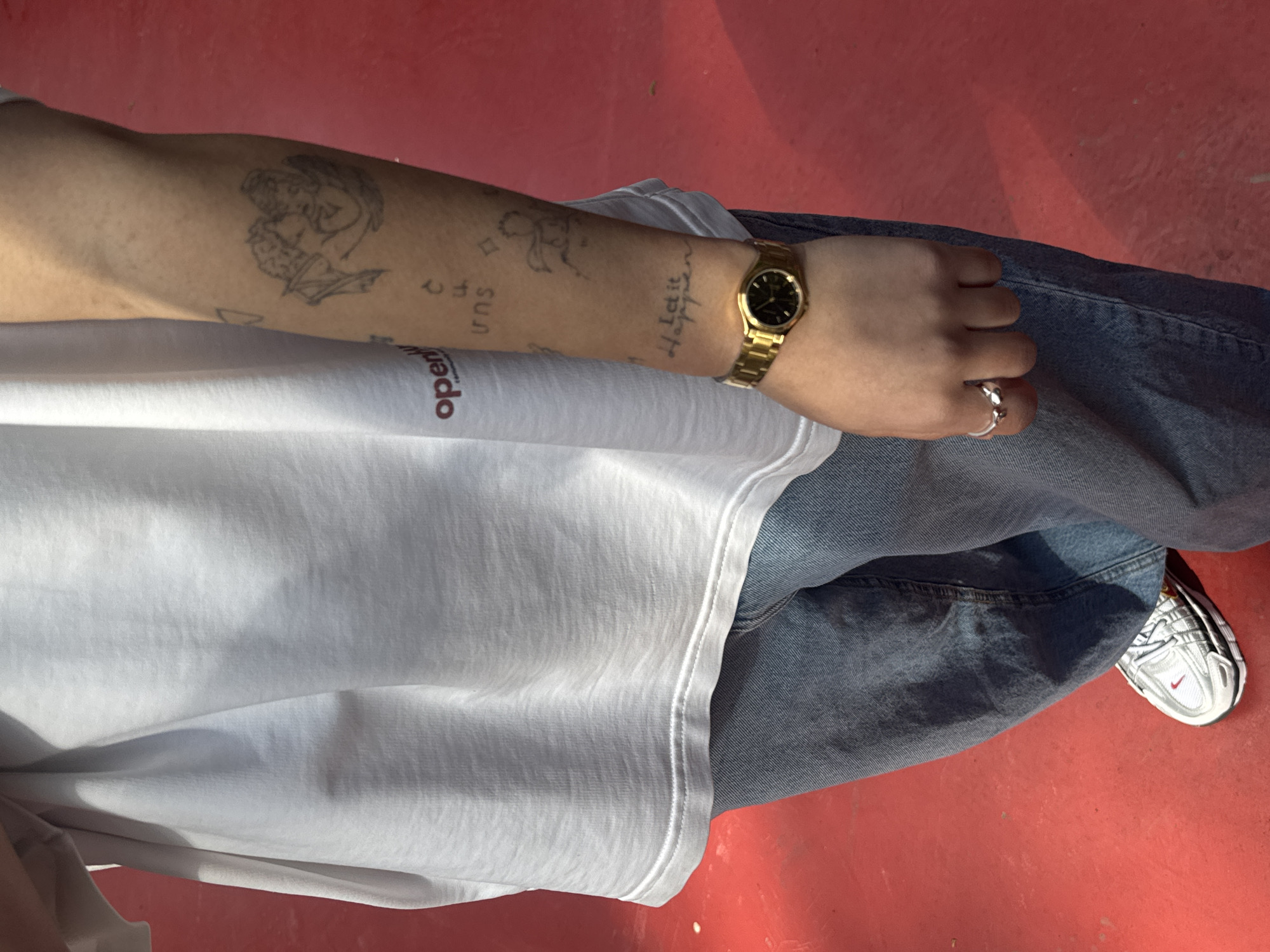The image depicts a person from a top-down perspective, dressed in casual clothing. The person is wearing a white T-shirt with the word "open" visible on the shirt. The person is wearing blue jeans. A gold wristwatch is on the left wrist. The person also has multiple tattoos visible on the left arm, including what appears to be an angel, the word "sun," and the phrase "Let It Happen." The person wears a silver ring on a finger of the left hand. The right foot, shod in a white Nike shoe with a red Swoosh, is visible. The setting appears to be outdoors with a red surface beneath. There is no evidence to confirm that this image was taken in Yerevan, Armenia.