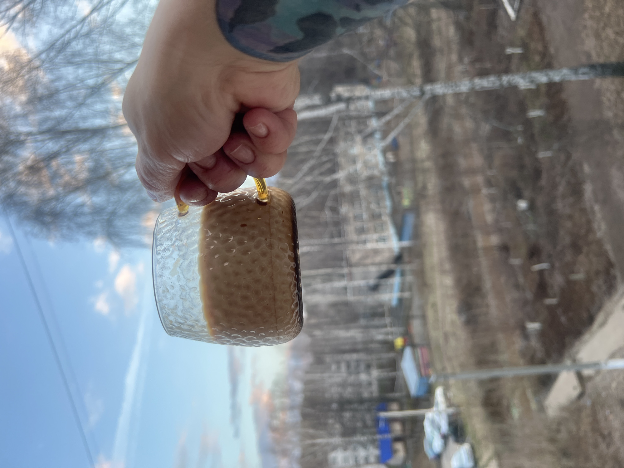 A hand, featuring a tattoo on the wrist, holds a glass mug with a yellow handle that is half full of a light brown liquid. The textured glass mug is centered in the image. The backdrop, blurred, shows a cityscape with buildings and trees, suggestive of an urban or suburban environment.
Based on the presence of Russian lettering and signage, and the request to identify the location as Chaikovsky, Russia, it's likely the picture was taken in this location.