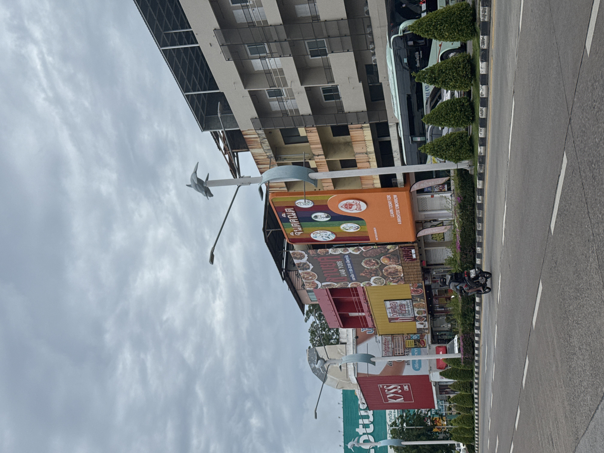 The image depicts a street scene in Pattaya City, Thailand, under an overcast sky. The foreground shows a multi-lane asphalt road with white dashed lane markings. In the midground, there is a paved sidewalk area with a row of manicured, conical evergreen trees and a low hedge. Beyond the trees, a variety of buildings and advertisements are visible.
On the left, a large green billboard with white lettering that appears to read "lotus" is present. Adjacent to it are several colorful buildings, including a bright red structure with "KYSS" in white lettering and "CLINIC" below. Next to the red building is a yellow building with "Bombay" and "Pines" written on it. An orange billboard with Thai text and images of food is also prominent. On the right side of the frame, a multi-story building with beige and grey facades, featuring balconies and dark-paneled windows, dominates the background. A teal and white bus is parked in front of this building.
Several people and vehicles are present. A person on a motorcycle is riding on the road in the right lane, facing away from the camera. A silver car is visible parked behind the trees. Two streetlights with decorative dolphin sculptures at the top are positioned along the sidewalk. The overall impression is a commercial street with shops and businesses lining the road.