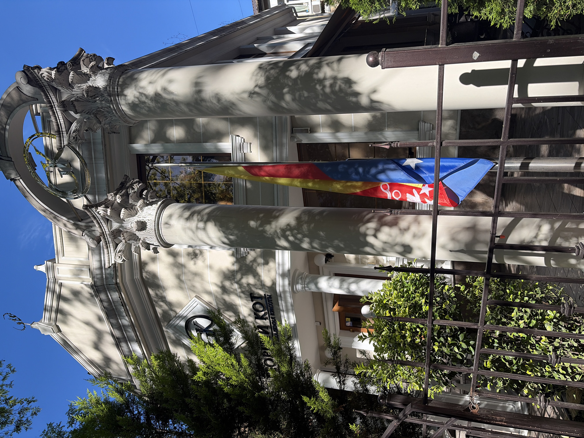 The image displays the exterior of a building in Sochi, Russia, bathed in bright sunlight under a clear blue sky. The building exhibits classical architectural elements, featuring two prominent columns with ornate, Corinthian-style capitals and a decorative arch-like structure above. Strong shadows from nearby trees and architectural details are cast across the facade and columns, indicating a sunny day.
A flag is prominently displayed, hanging vertically from the building. It features horizontal stripes of red, yellow, and blue, with a white five-pointed star set within a blue triangle on the hoist side, consistent with the design of the Catalan Estelada flag.
In the foreground, a dark metal fence with decorative finials partially obstructs the view. Lush green foliage, including bushes and trees, is visible both in front of the fence and along the building's base.
A sign on the building's facade clearly displays text in Cyrillic: "БЕЛЫЙ КОТ САЛОН КРАСОТЫ" (BELY KOT SALON KRASOTY), which translates to "White Cat Beauty Salon." No individuals or specific activities are depicted, portraying a serene, static street scene.