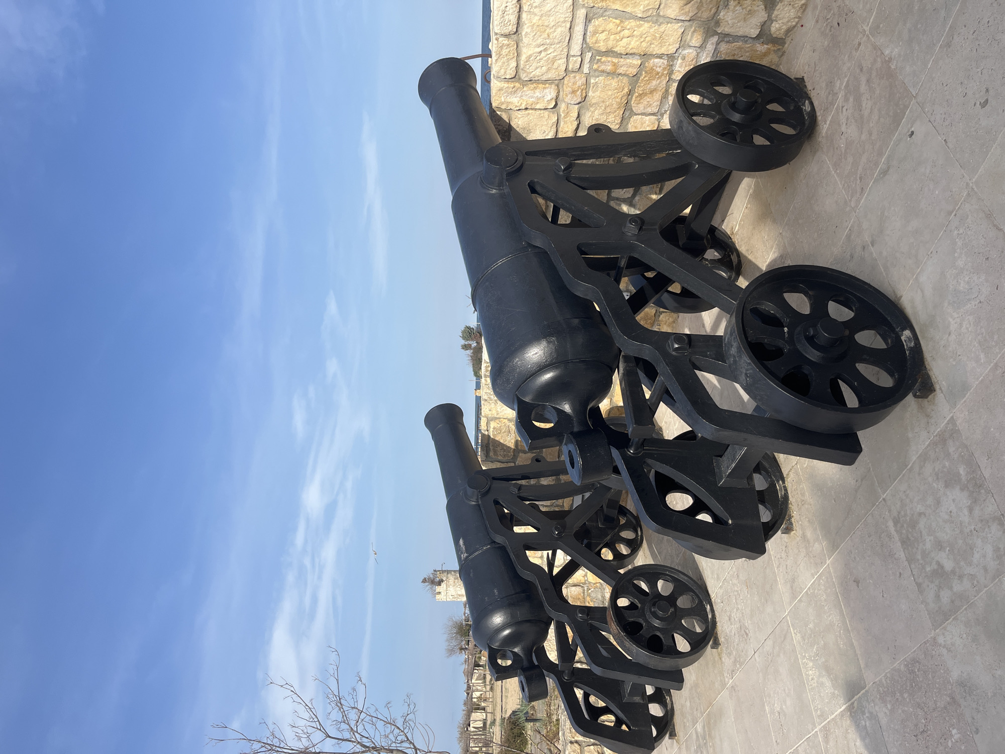 The image features two black cannons positioned on a stone surface, possibly a platform or wall. The cannons are mounted on wheeled carriages, also painted black. Behind the cannons, there is a stone wall, and beyond that, a view of the sea and sky is visible. The sky is blue with some clouds. The cannons appear to be historical artifacts, and the location seems to be a coastal area. It is likely located in Varna, Bulgaria.