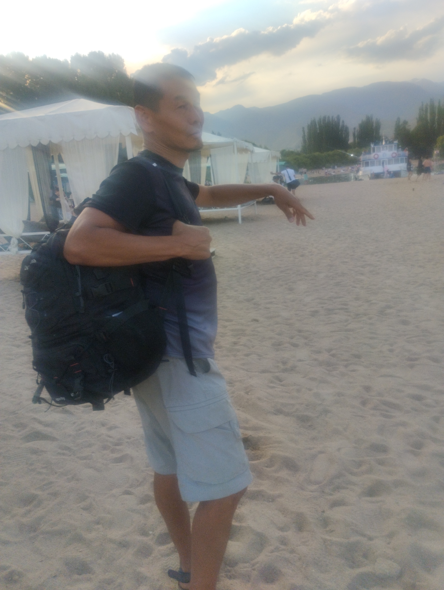 An adult male, appearing to be of Asian descent, stands on a sandy beach, wearing a black t-shirt and light-colored shorts. He has a large black backpack slung over his left shoulder. His right arm is extended, with his hand pointing towards the right side of the frame. In the background, there are white tents, trees, mountains, and a white boat with a red stripe on the water. The overall scene suggests a recreational area on a body of water, likely a lake, with mountains in the distance. The lighting indicates it is late afternoon or early evening, with soft sunlight filtering through clouds. This location is consistent with the shores of Lake Issyk-Kul in Cholpon-Ata, Kyrgyzstan, a popular tourist destination known for its beaches, tents, and mountain backdrop.