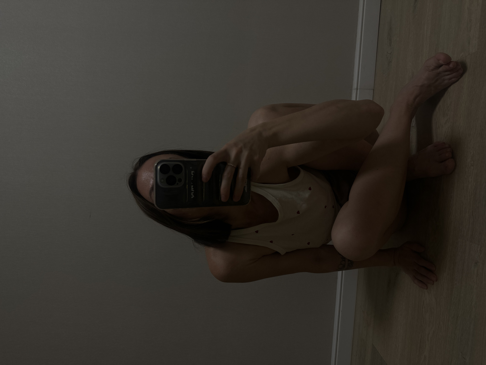 A woman is crouched on a wooden floor, taking a selfie with her phone, which obscures her face. She is wearing a light-colored tank top with small red hearts. Her left arm is bent, and her hand rests on her knee. Her right arm is extended, holding the phone. A ring is visible on her left ring finger. Her legs are bent, and her feet are resting on the floor. A tattoo is visible on her left arm. The background is a plain, dark wall. The scene appears to be indoors. The image does not contain any information that would indicate its location in Votkinsk, Russia.