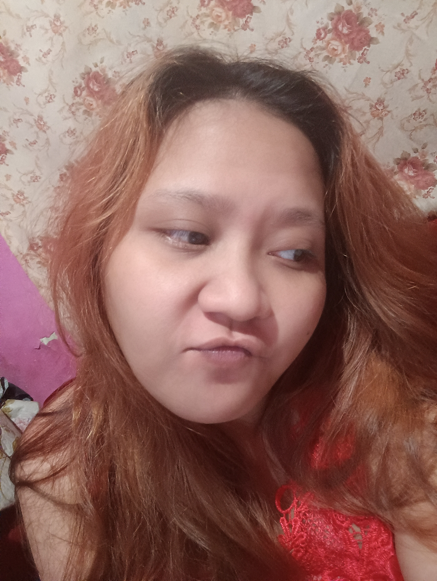 A close-up, selfie-style photo shows a young woman with auburn hair against a floral-patterned wallpaper. She is looking to her right with her lips slightly puckered. A portion of a pink surface is visible on the left side of the frame, and she is wearing a red garment with lace detailing on the right. The image was taken indoors, likely in a residential setting in Depok, Indonesia, as suggested by the background and overall style.