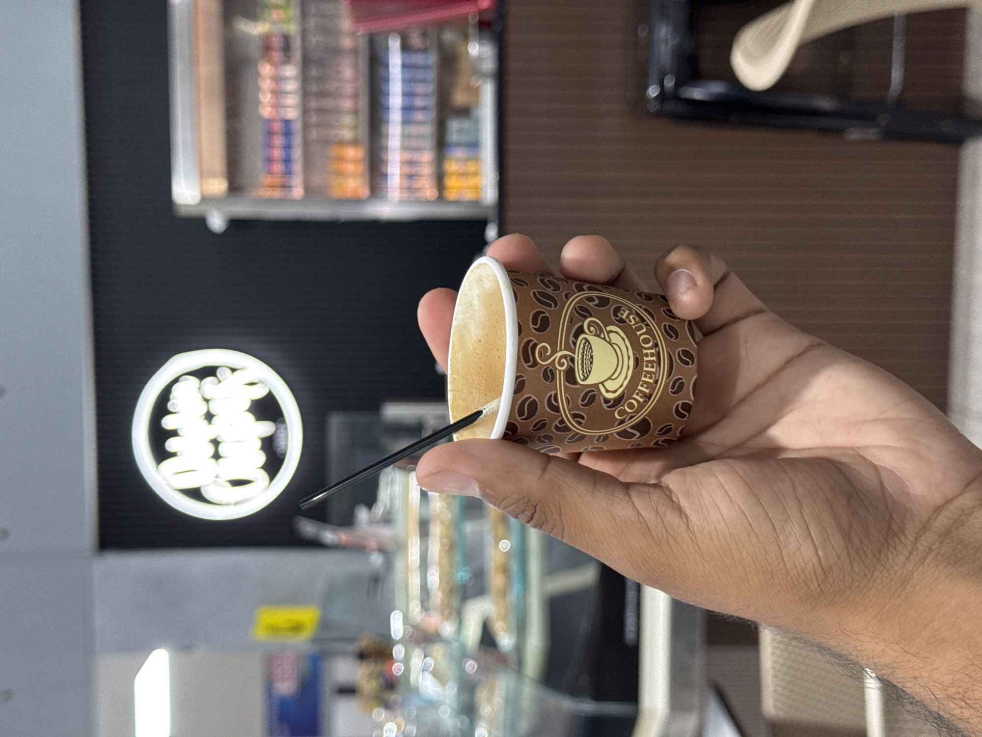 A close-up photograph shows a hand holding a small, brown paper coffee cup labeled "COFFEEHOUSE." A black stirrer is inserted into the cup. The background features a black and white wall display with a neon-lit Chinese character sign. Below the hand, out-of-focus items are seen on a glass shelf. The location is likely Porlamar, Venezuela.