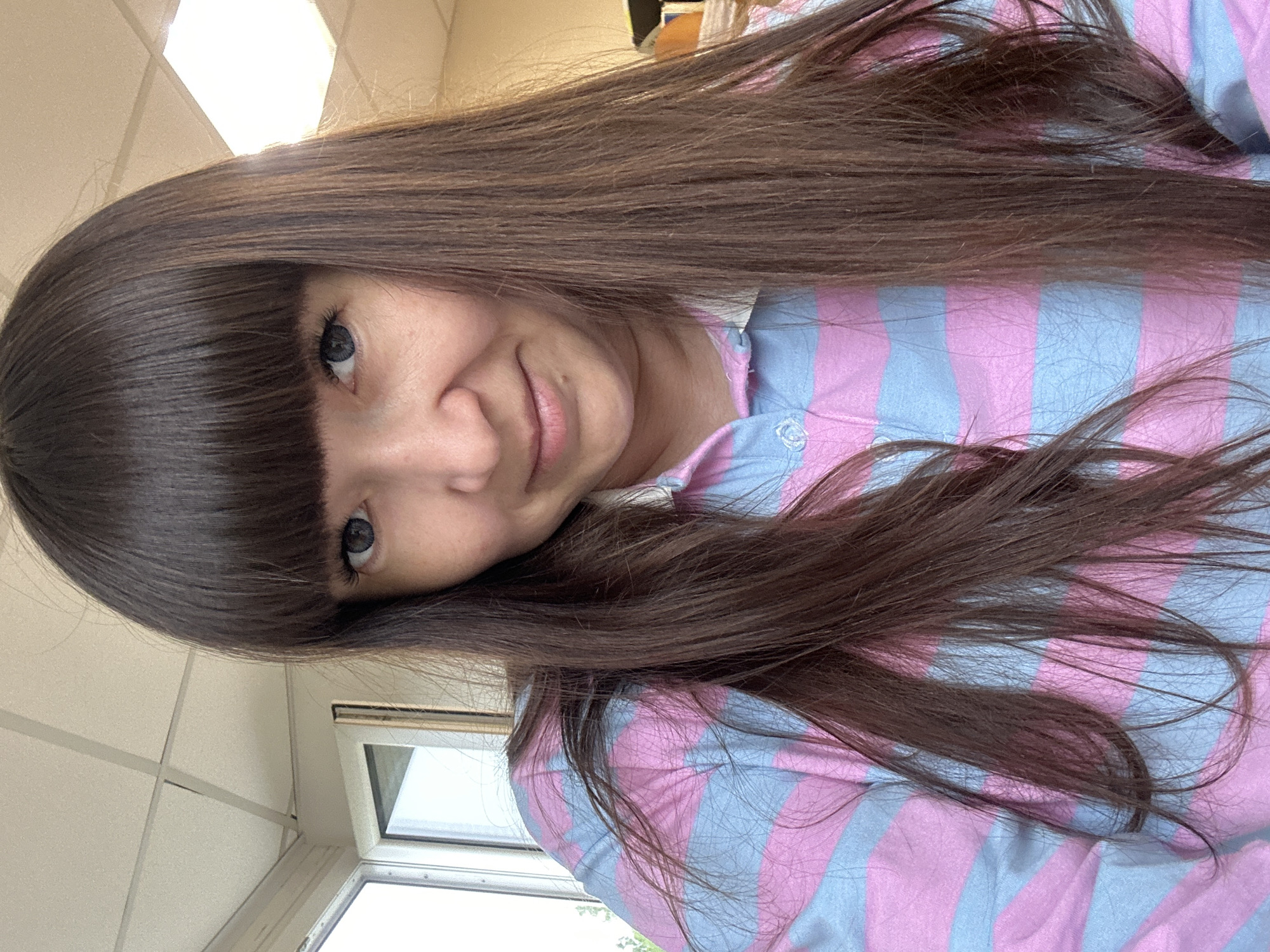 A medium close-up shot shows a woman with long, dark brown hair with bangs. She is wearing a pink and blue striped long-sleeved shirt with a collar. The woman is looking at the camera with a slight smile. The background appears to be an indoor environment with a white ceiling, recessed lighting, and a window with a view of trees. There is no information available to specify the location in the city of Chaikovsky, Russia.