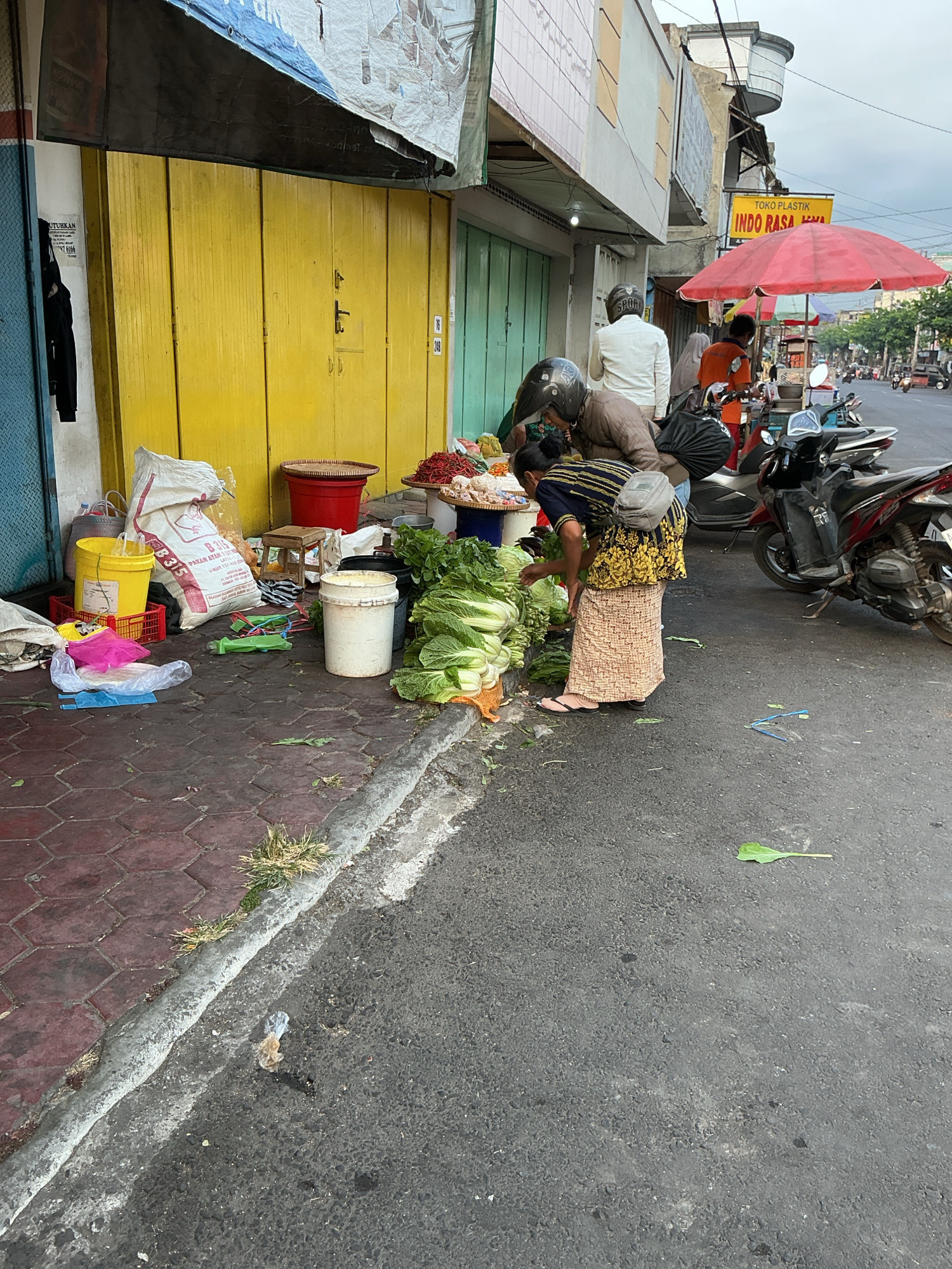 This image depicts a street scene in Magelang, Indonesia, likely in a market area. In the foreground, a woman wearing a dark blue and yellow patterned top and a patterned skirt is bent over a large pile of leafy green vegetables, possibly cabbage. She has a light gray bag slung over her right shoulder. Next to her, another person wearing a brown jacket and a helmet is also looking at the vegetables. Behind them, several motorcycles are parked on the street.
To the left, a yellow shuttered storefront is visible, with various items piled on the ground including bags, buckets, and plastic bags. A large white sack with red lettering is propped against the wall. In the background, a red umbrella shades a food stall, and more motorcycles and vehicles are seen on the street, indicating a bustling urban environment. The pavement is made of reddish-brown hexagonal bricks, and the street is asphalt. The overall impression is of an active marketplace with vendors and shoppers.