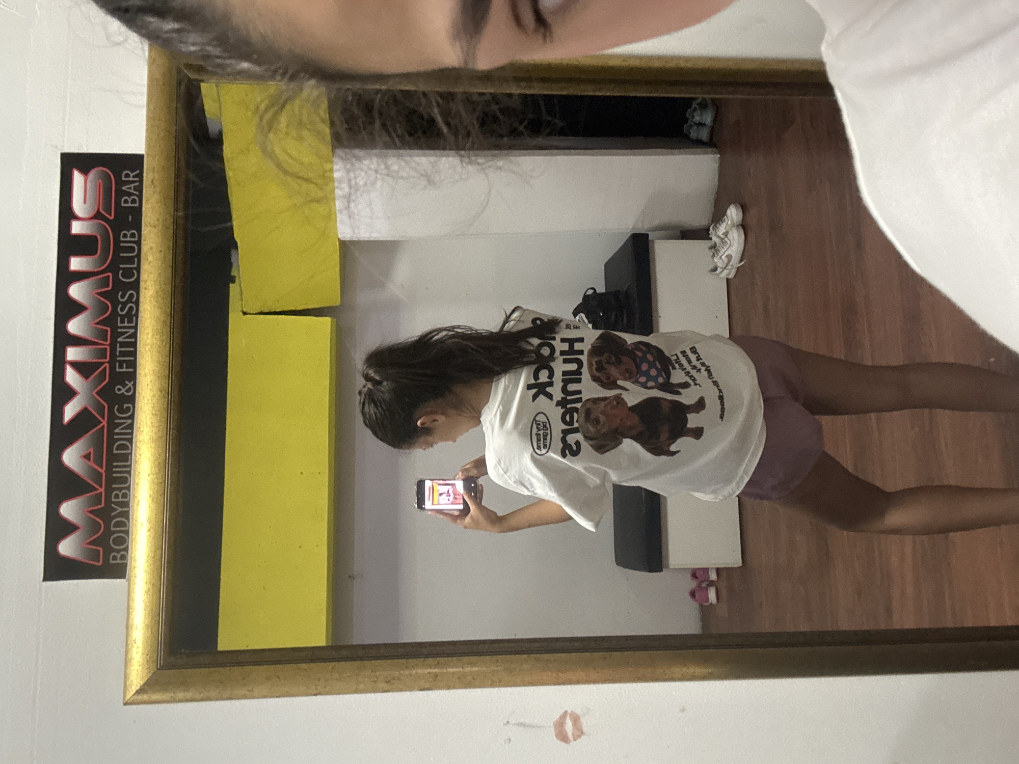 A reflection in a gold-framed mirror captures a young woman taking a photo of herself with her smartphone. The mirror hangs on a white wall in what appears to be a gym. Visible in the reflection are yellow padding, a black weight bench, and flooring resembling wood. The woman in the reflection is wearing a white t-shirt with a print of dachshunds and lilac shorts. A "MAXIMUS BODYBUILDING & FITNESS CLUB - BAR" sign is affixed to the wall adjacent to the mirror. A smudge resembling a lipstick mark is visible below the frame. The location is indicated as the city of Bar, Montenegro.