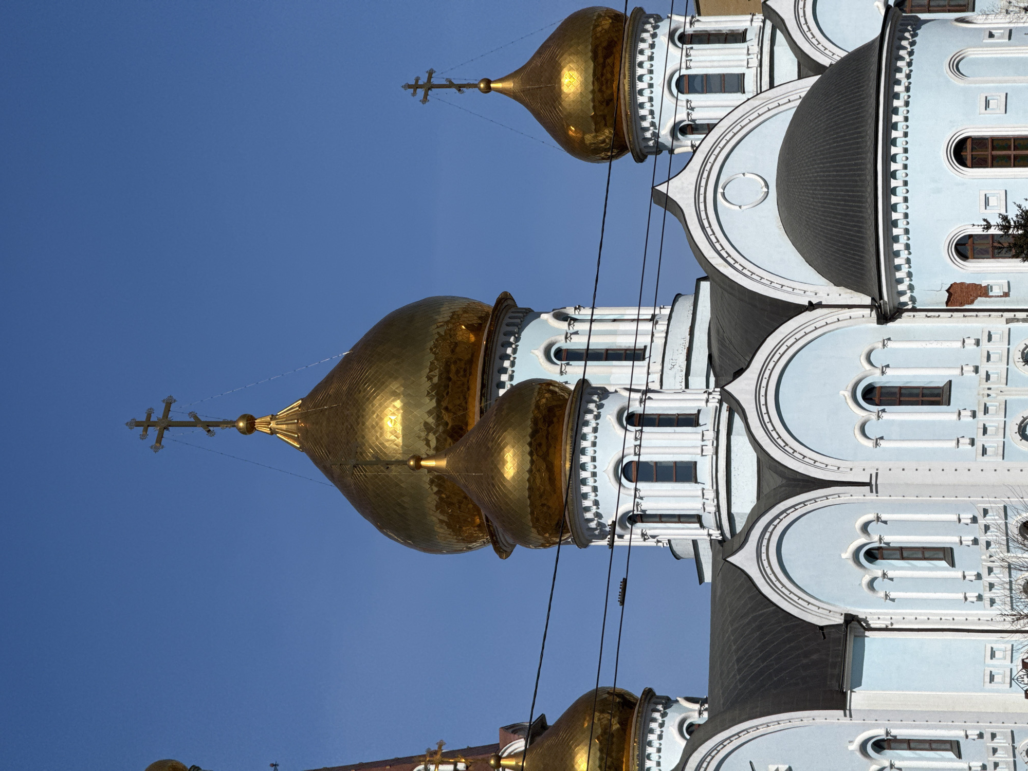 The image shows the Church of the Kazan Icon of the Mother of God in Chita, Russia. The building has golden domes with crosses atop them. The walls of the church are light blue. The background is a clear, bright blue sky. There are power lines visible in the image.