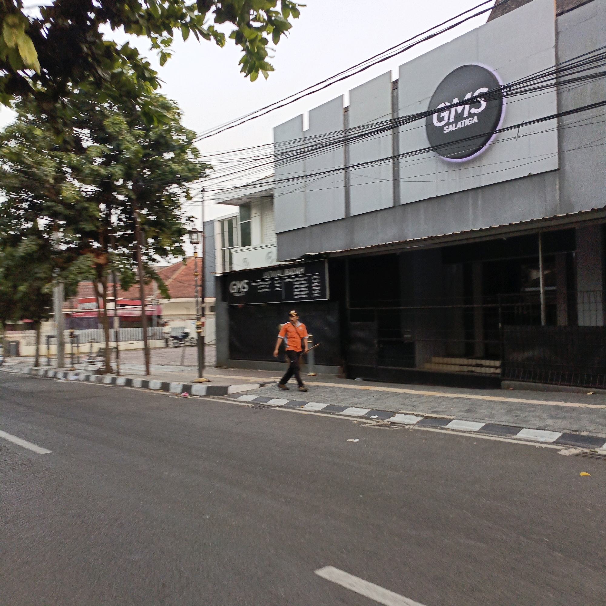The image shows a street scene in Salatiga, Indonesia. A man wearing an orange shirt and dark pants is walking on the sidewalk. He is carrying a long stick or pole. Behind him is a building with a large circular logo that says "GMS SALATIGA". The building appears to be a commercial establishment. Trees and power lines are visible in the background, suggesting an urban environment. The street is paved and has a pedestrian crossing with black and white markings. The overall atmosphere is that of a typical day in an Indonesian city.