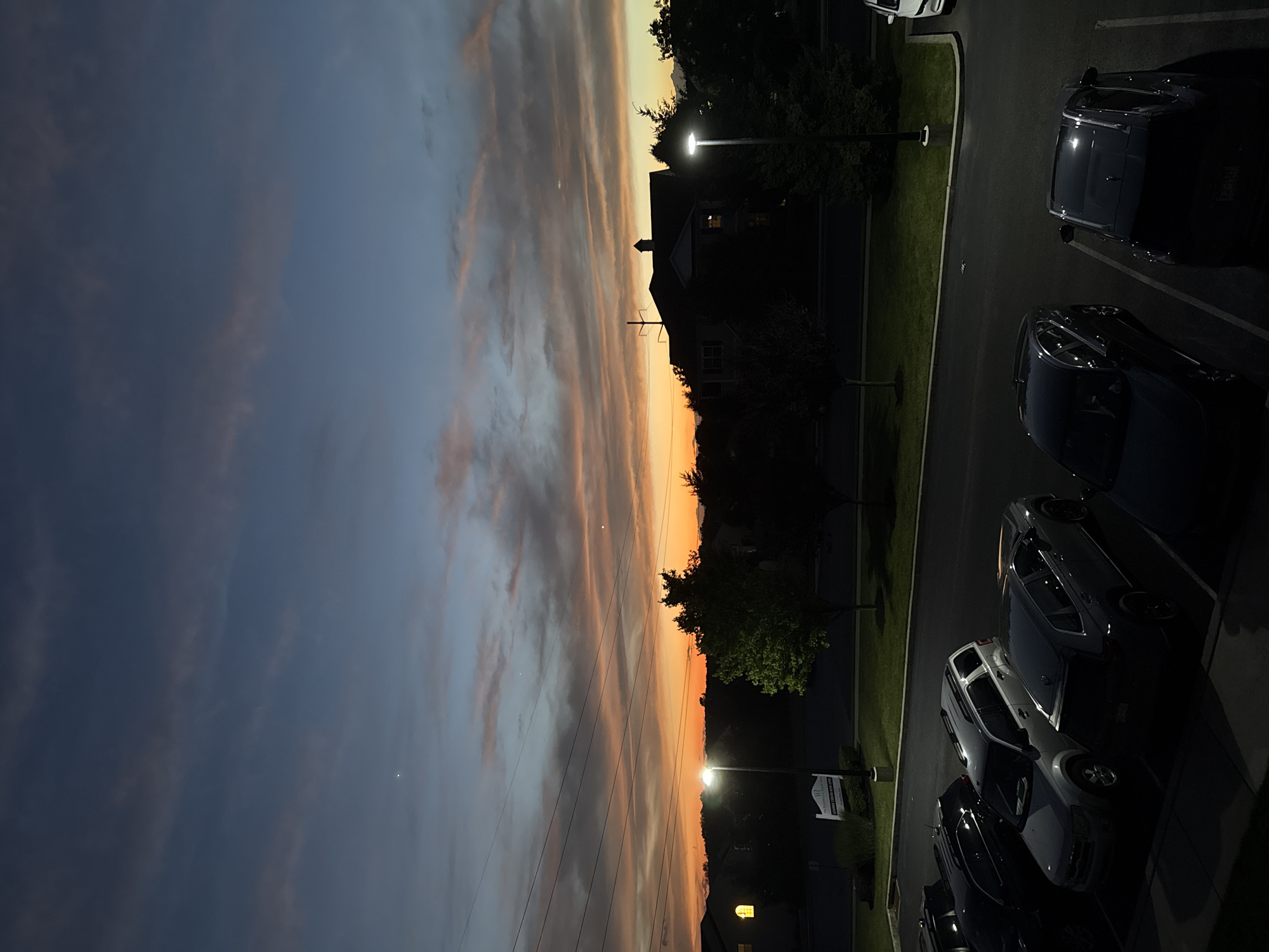 The image depicts a twilight scene over a residential area, likely a parking lot, in Allen Township, United States of America. The sky is painted with hues of deep blue, orange, and pink, indicating the sun has recently set. Wispy clouds stretch across the horizon. In the foreground, several cars are parked in designated spots, their forms partially obscured by the low light. A streetlamp illuminates a portion of the parking lot, casting long shadows. Behind the parking lot, silhouetted houses and trees are visible. The overall impression is one of quiet evening transitioning into night in a suburban or apartment complex setting.