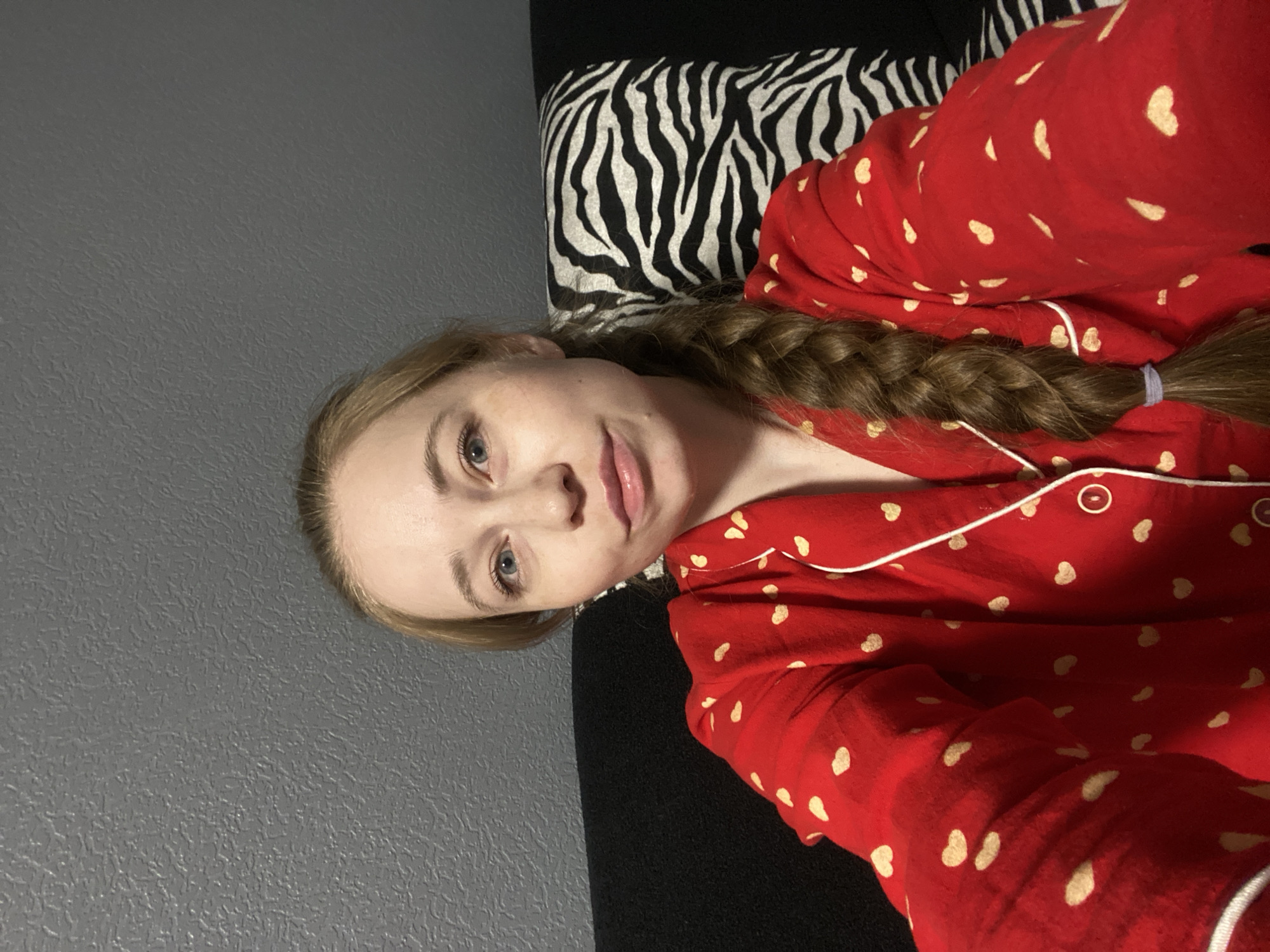 A young woman is captured in a selfie, positioned as if lying down or reclining. Her head is to the left of the frame, with her body extending to the right. She has light skin, blue eyes, and light brown or blonde hair styled in a long braid that drapes over her shoulder and chest. A light purple hair tie secures the end of her braid. She wears red pajamas featuring a pattern of small, light-colored heart shapes and white piping. Her expression is calm, with a subtle smile.
The background consists of a textured dark grey wall to the left, behind her head. Behind her upper body and shoulder, a plain black surface is visible, possibly a couch or headboard. Further to the right and behind her lower body is a pillow with a distinctive black and white zebra print pattern. The indoor setting, along with her pajama attire, suggests the activity is taking place in the evening or night, likely in a bedroom or living room. The image provides no specific visual cues for the given location context of Saint Petersburg, Russia. There is no visible text in the media file.