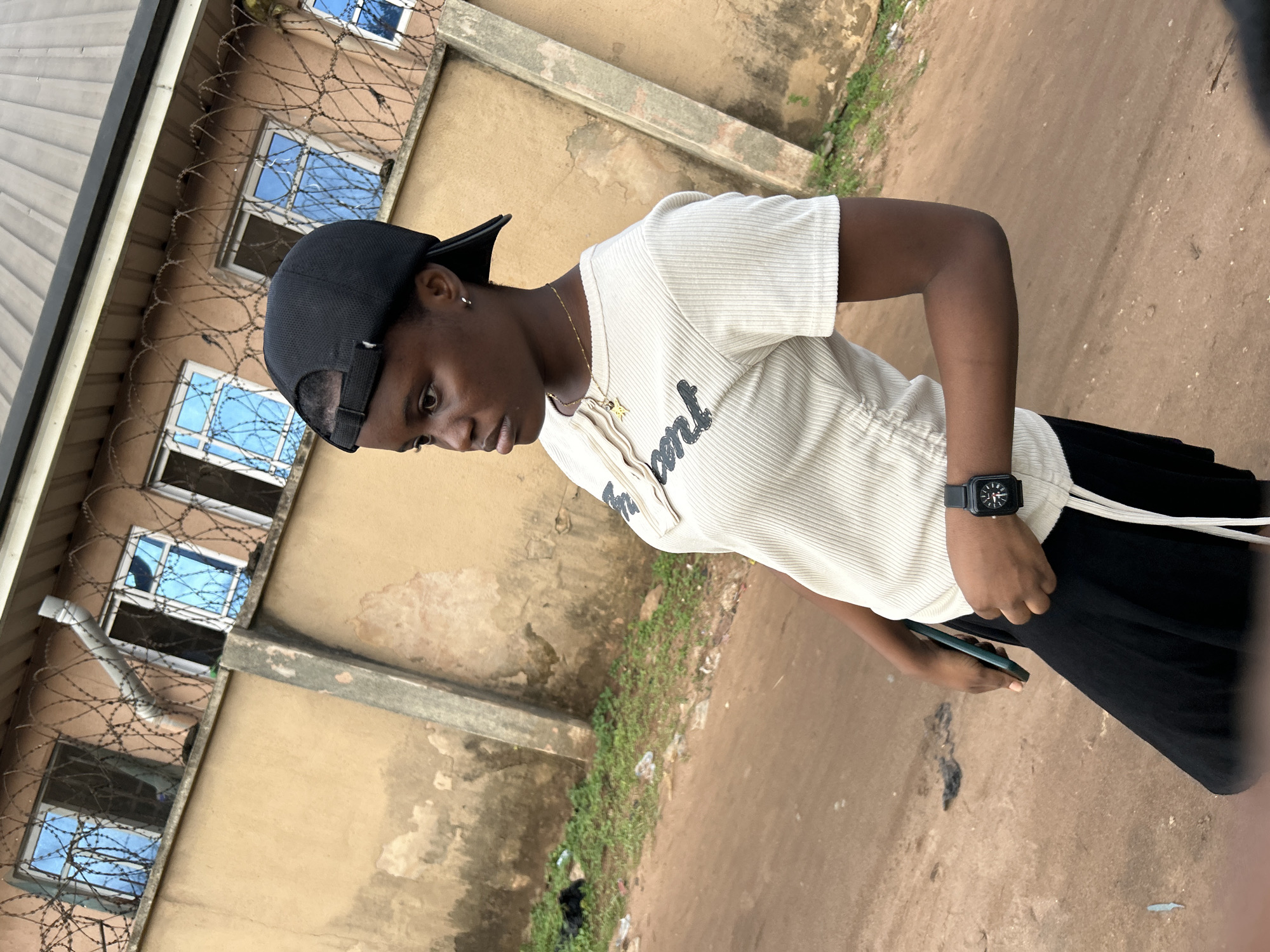 A young Black female stands in front of a building with a tan, textured wall and multiple windows with bright blue glass, located in the city of Ihiagwa, Nigeria. Barbed wire is stretched horizontally across the top of the wall. The female wears a black baseball cap, a light-colored ribbed t-shirt with a drawstring detail at the waist, and black pants. She has a gold necklace with a star pendant and a black wristwatch. She is holding a dark-colored object, possibly a phone, in her left hand. The ground in front of her is a dirt road with sparse green vegetation and scattered debris. The lighting suggests it is daytime.