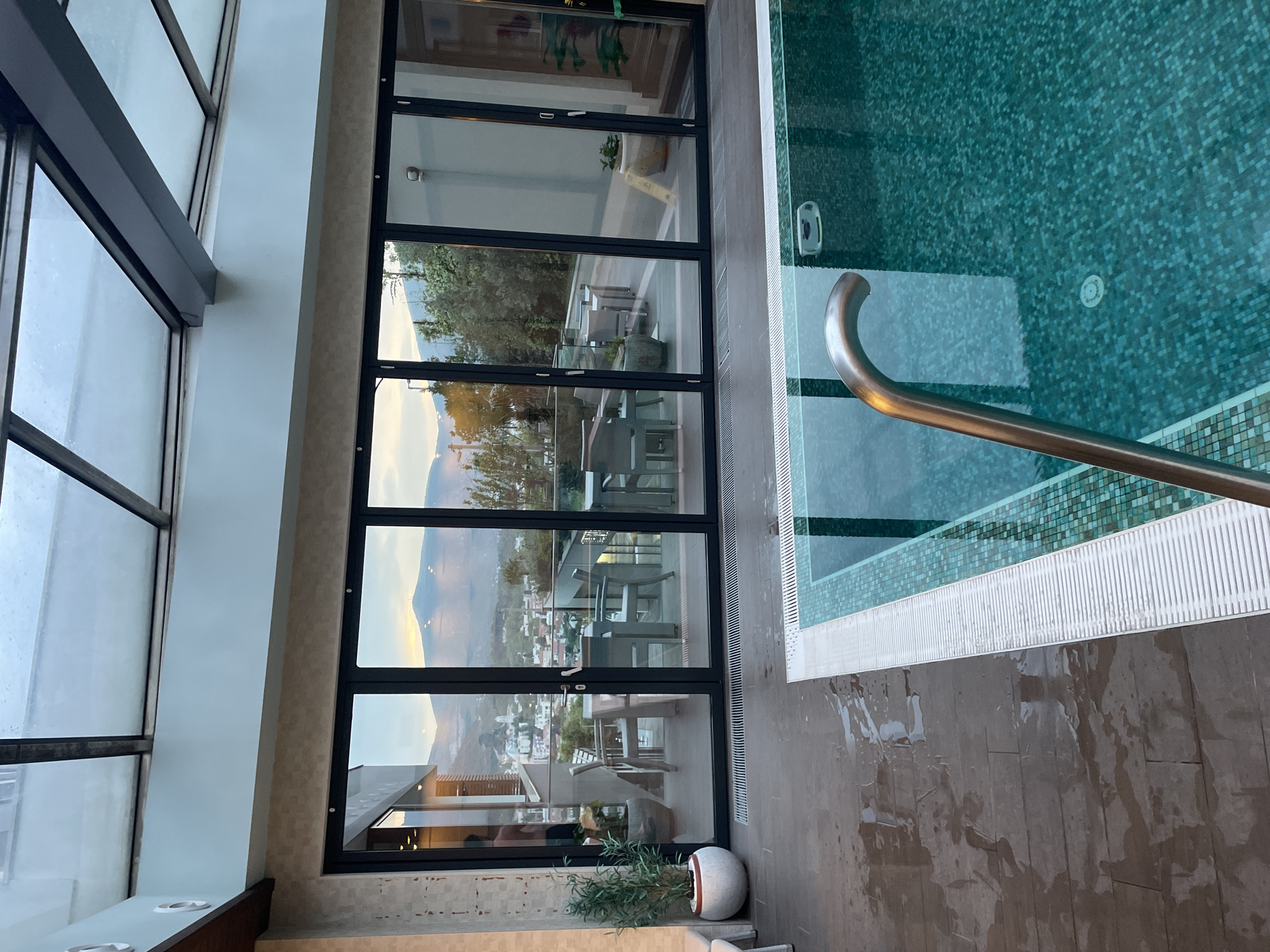 A high-angle interior shot depicts a tiled pool area and a window wall reflecting an outdoor scene, potentially in the city of Mostar, Bosnia and Herzegovina, based on the visible landscape. The pool, filled with turquoise water, features a stainless steel handrail. The area around the pool is tiled with dark brown material that appears wet. A large window wall with black frames separates the pool area from the exterior. The reflection in the glass shows tables and chairs on a terrace, a hillside, and a cityscape in the distance. A potted plant sits near the base of the window wall. The window wall is flanked by a multi-paneled glass structure, suggesting a high-ceilinged or sunroom-like area. The lighting appears to be a mix of natural light and artificial lighting.