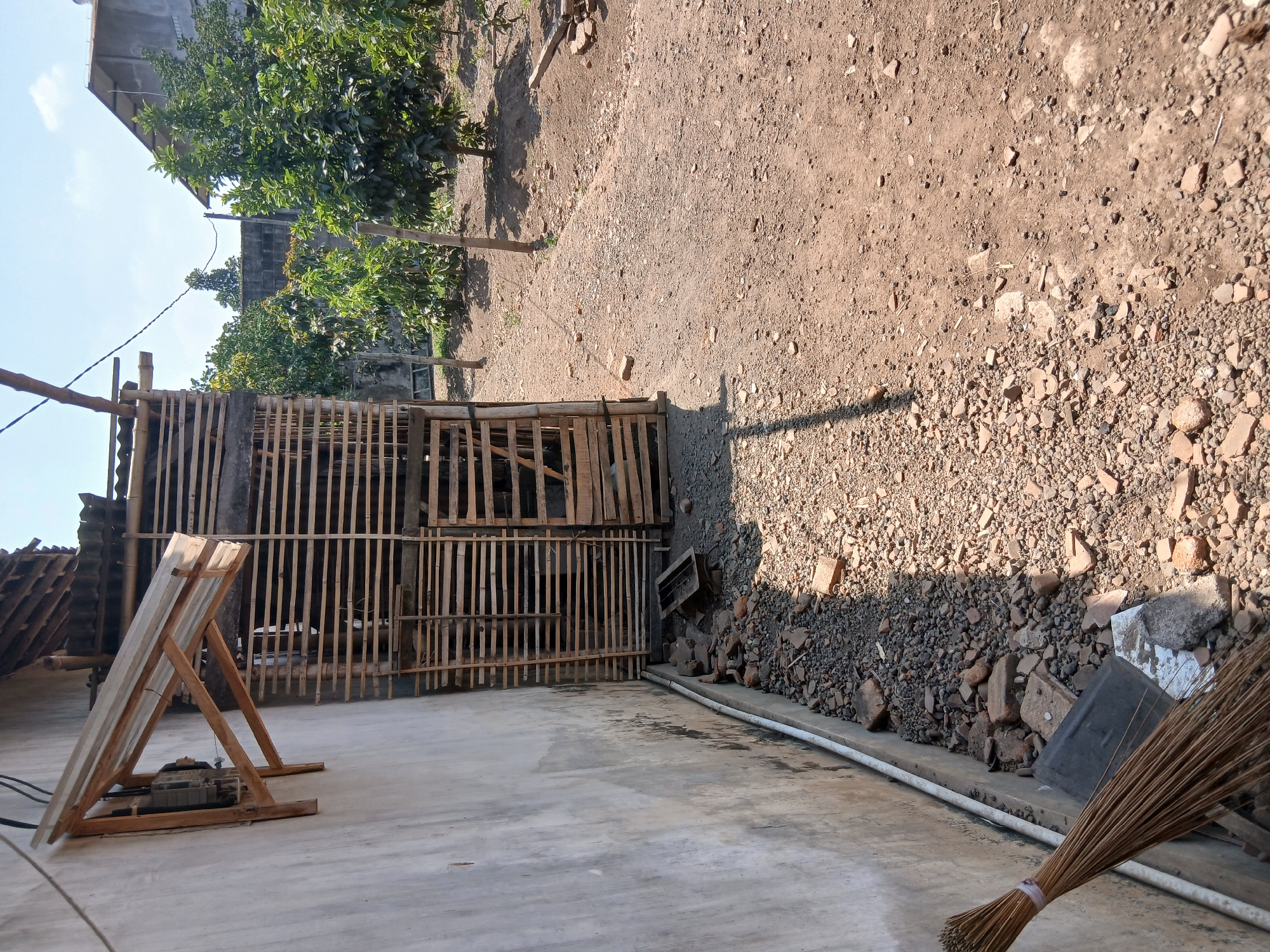 The image depicts an outdoor scene on a sunny day, likely in a rural or semi-rural area of Tenggarang, Indonesia. In the foreground, a broom made of dried palm fronds leans against a rough, light-colored concrete wall. A white conduit pipe runs along the base of the wall. The ground is covered with a mixture of small stones, gravel, and broken pieces of reddish-brown brick or tile. To the right of the broom, a dark gray rectangular object and some other debris are scattered on the ground.
Adjacent to the wall is a rustic structure constructed from horizontal wooden slats, resembling a cage or enclosure. This structure has a corrugated metal roof supported by wooden beams. The slats are closely spaced, with some openings visible. A wooden plank leans against the side of this structure.
Further into the background, the ground transitions to dry, reddish-brown soil with scattered stones and small plants. A line of wooden posts marks a boundary. Behind these posts, several trees with dense green foliage are visible, suggesting an orchard or garden area. In the far background, parts of buildings are discernible, one with a gray, textured facade and a dark roof, and another, more distant structure with a lighter colored roof. The sky is blue with scattered white clouds. The overall impression is one of an informal, utilitarian outdoor space.