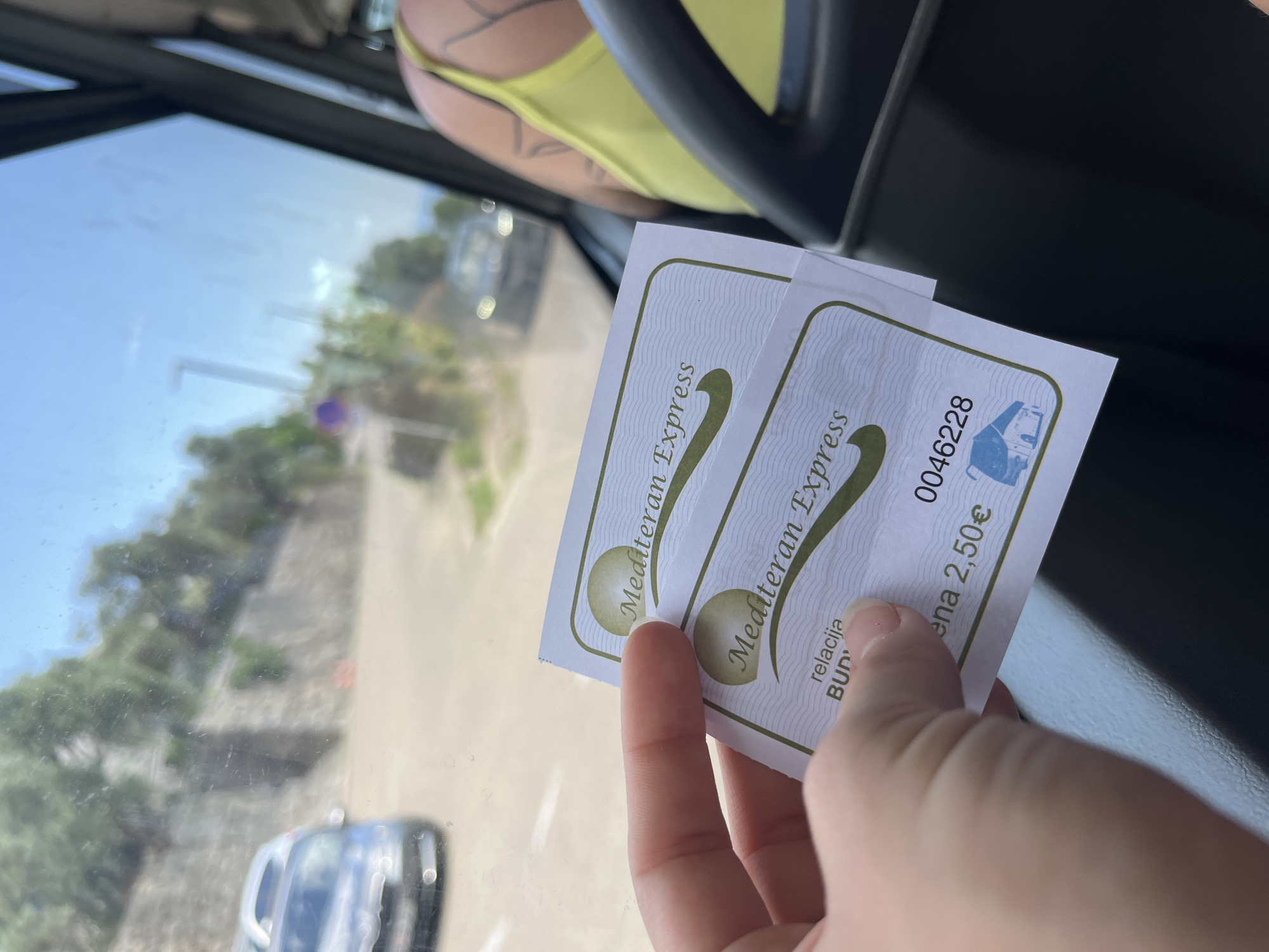 A passenger inside a vehicle holds two "Mediteran Express" bus tickets, priced at 2.50€ each, indicating a trip originating in Budva, Montenegro. Outside the window, the blurred scenery includes greenery, a retaining wall, and vehicles, suggesting a roadside view in Sveti Stefan, Montenegro. The passenger is wearing a yellow top and has a tattoo on their shoulder.