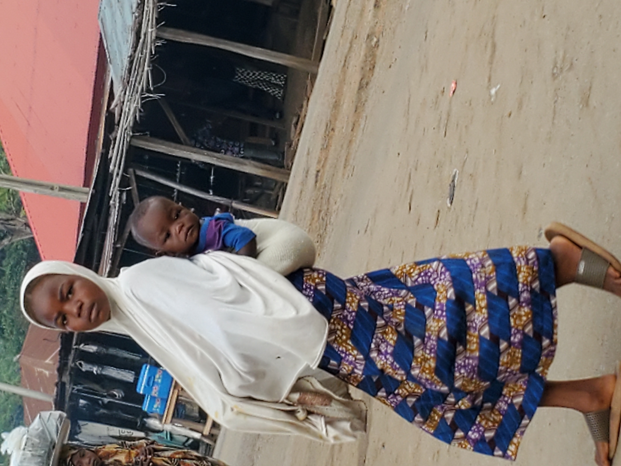 A woman, dressed in a white headscarf and a patterned blue and purple skirt, carries a baby on her back. They are standing on a dirt surface in what appears to be a populated area, possibly Mararaba, Nigeria. There are buildings with corrugated metal roofs in the background.