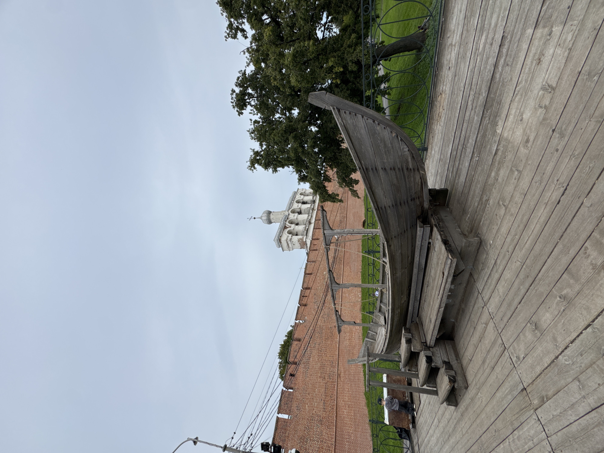 This is a wide-angle, outdoor, daytime photograph taken in Veliky Novgorod, Russia, likely in the vicinity of the Veliky Novgorod Kremlin. In the foreground, a weathered wooden boat, possibly a historical replica, rests on a wooden platform. The boat has a distinct curved hull and a rudimentary wooden canopy structure. Wooden steps lead up to the boat.
In the mid-ground, a red brick wall, characteristic of fortress architecture, stretches horizontally across the frame. The wall has battlements along its top. Behind the wall, a green lawn is visible, separated from the wooden platform by a decorative metal fence. A large, leafy tree stands to the right of the frame, partially obscuring a portion of the green space.
In the background, a white bell tower with multiple arches and a cross on top rises above the brick wall, a prominent architectural feature of the city. The sky is overcast and pale gray.
To the left of the boat, a young child wearing a gray hooded jacket and dark pants stands with their back to the camera, looking towards a dark object on the wooden platform. Overhead, a complex network of electrical wires and poles is visible against the sky, suggesting the presence of urban infrastructure. The overall scene evokes a sense of historical significance and public space within a city.