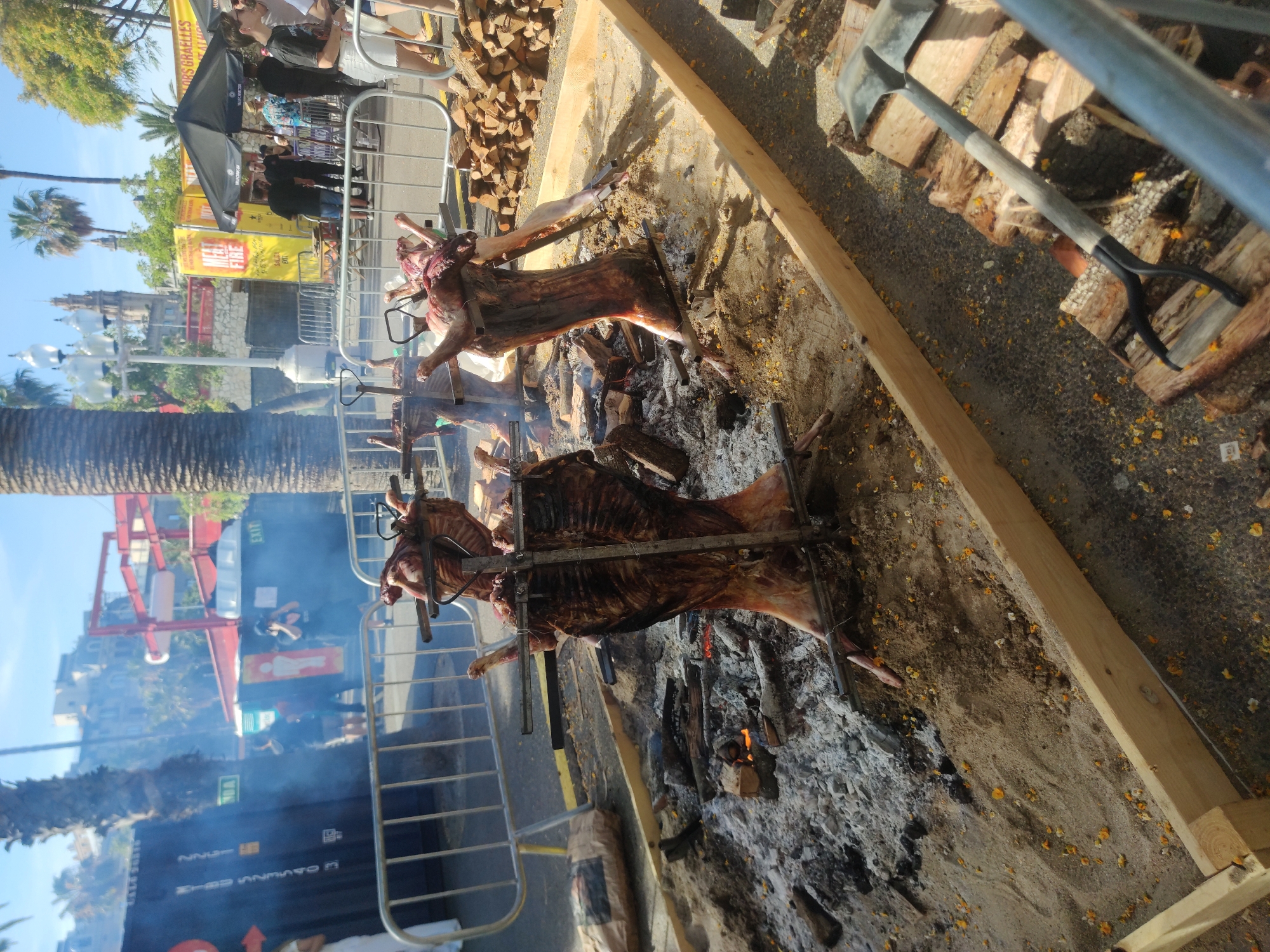 In an outdoor setting, possibly a street market or festival, two whole pigs are being roasted on spits over an open fire. The pigs are suspended on metal frames, with one pig positioned more towards the foreground and another further back. The fire is built within a shallow sand pit, surrounded by a wooden frame. Ash and burning embers are visible around the base of the roasting pigs. In the background, a crowd of people are gathered, some standing under tents and others near food stalls. Smoke billows from the cooking area, obscuring some of the background activity. A stack of chopped firewood is visible to the right of the roasting pits, along with a shovel. The scene appears to be under a clear sky with sunlight illuminating the area. The location is described as Unknown, Unknown.