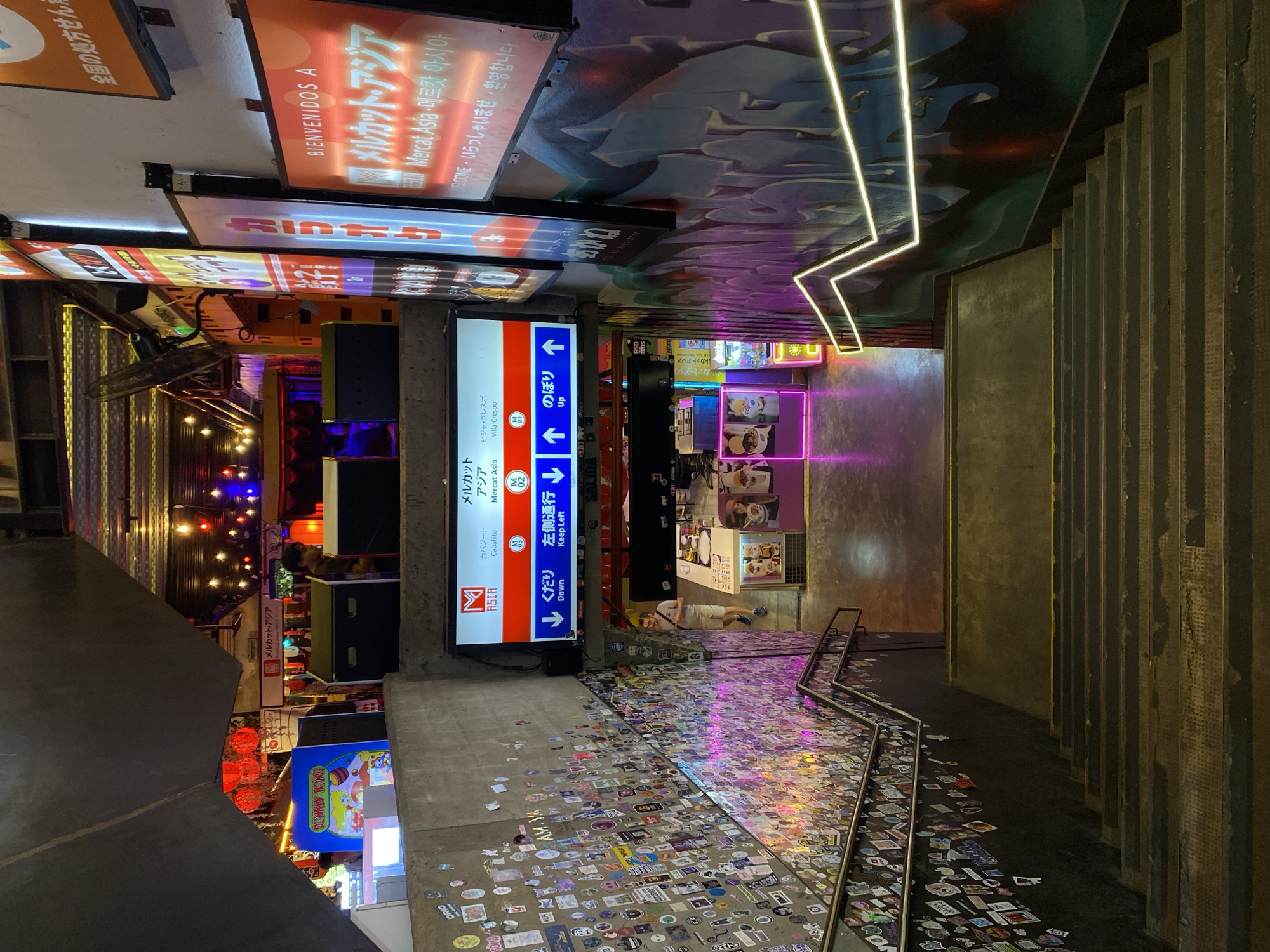 The image shows a stairway and hallway within a vibrant commercial district, possibly a food court or marketplace, in Buenos Aires, Argentina. A sign in the center, indicates "Mercat Asia" in multiple languages, directing patrons up and down the stairway. The hallway walls are densely covered in stickers. Numerous signs above advertise various businesses, including "Mercat Asia" again, with the phrase "Bienvenidos A". The scene is visually stimulating with bright lights, neon signs, and a mix of colors. The stairway itself is well-lit and leads to an area further in the building. A person can be seen walking in the marketplace.