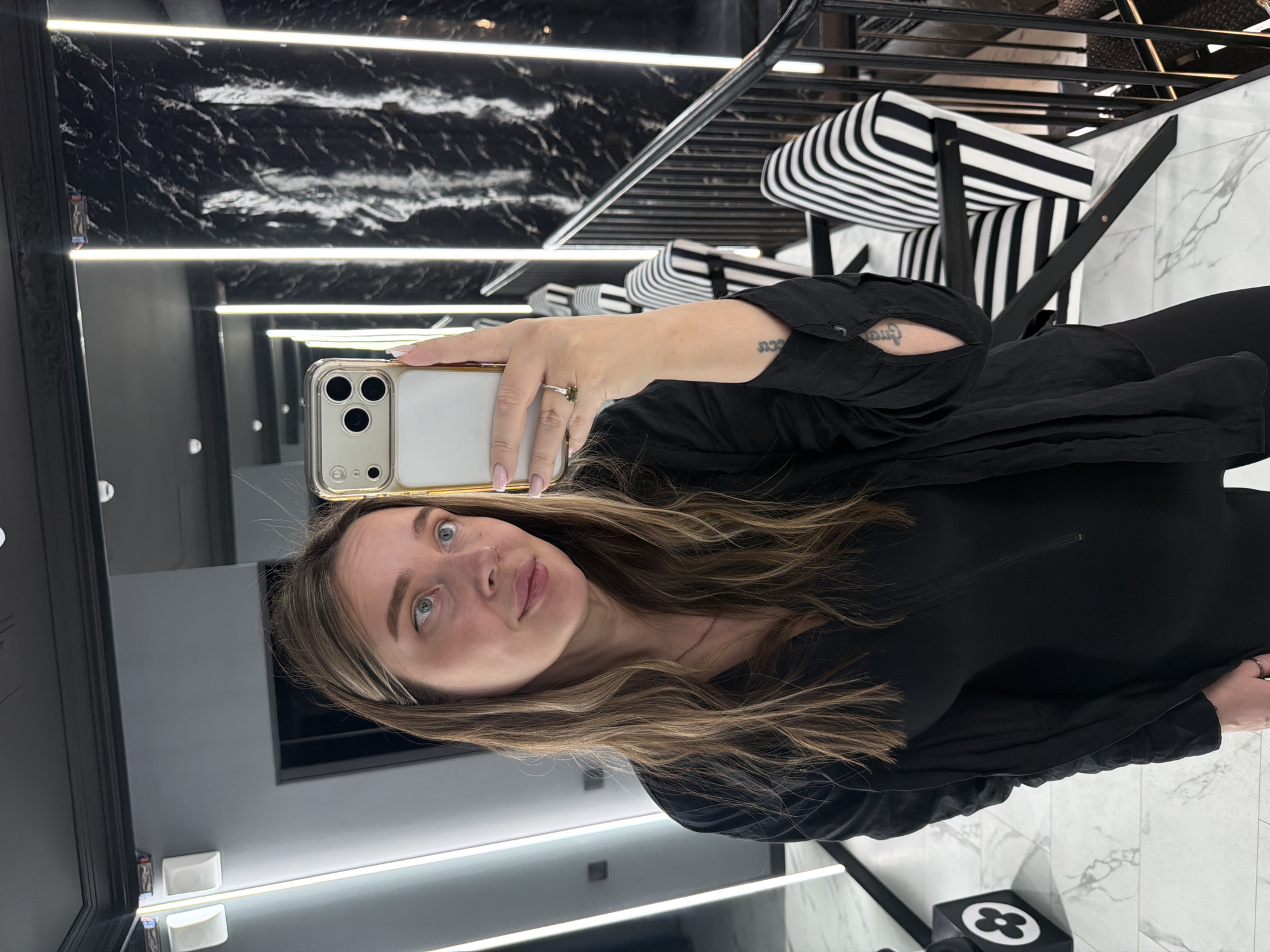 A young woman, likely in her 20s or 30s, captures a mirror selfie indoors, presumably in Moscow, Russia. She has long, wavy brown hair with lighter highlights and light-colored eyes, looking directly into the camera with a subtle smile or smirk. She wears a black long-sleeved top or dress. Her left hand, adorned with a ring featuring a green stone and manicured nails with light pink polish, holds a light-colored smartphone with a gold-trimmed camera module, characteristic of an iPhone, which is visible in the reflection. A small tattoo, appearing to be mirrored Hebrew script (possibly "כחב"), is visible on her inner left wrist.
The setting is a modern, stylish interior characterized by high-contrast black and white elements. The walls feature a dark, possibly black marble pattern, illuminated by multiple horizontal strip lights that create a sense of depth. In the background, black and white striped chairs, possibly lounge or dining chairs, add to the contemporary aesthetic. The floor is a light-colored marble pattern. A black cube with a white four-petal flower symbol, resembling the Louis Vuitton monogram, rests on the floor, enhancing the upscale ambiance of the room. The artificial lighting indicates the time of day is not discernible from natural light.