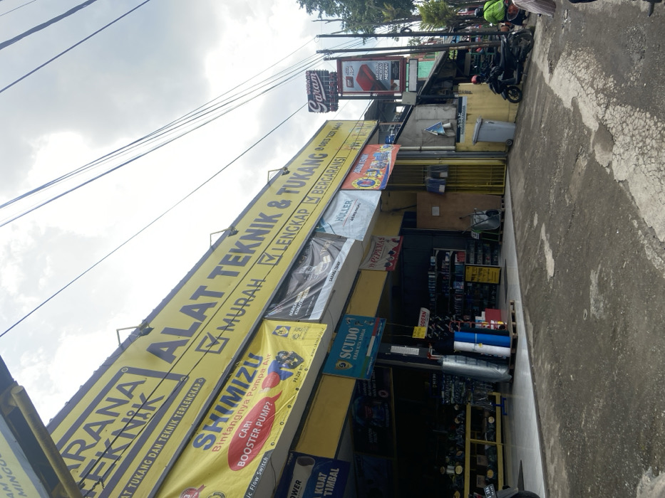 The image shows a store in Ciparay, Indonesia, located at latitude -7.029 and longitude 107.697. The store is a hardware store, selling a variety of goods, including paint, plumbing supplies, and tools. The store has a yellow awning with the words "Alat Teknik & Tukang" written on it. There are several other signs in front of the store, advertising different brands of products. There are also several motorbikes parked in front of the store, and a man is walking towards the camera. The environment is urban and is a street with a sidewalk in front of the shop. The day is cloudy, and there are power lines overhead. The sidewalk is paved and there is a trash can on the sidewalk in front of the store. The man is wearing a green shirt and has his back turned to the camera. The motorbikes are parked in front of the store on the sidewalk. The power lines are strung along the street. The image is taken at a low angle.